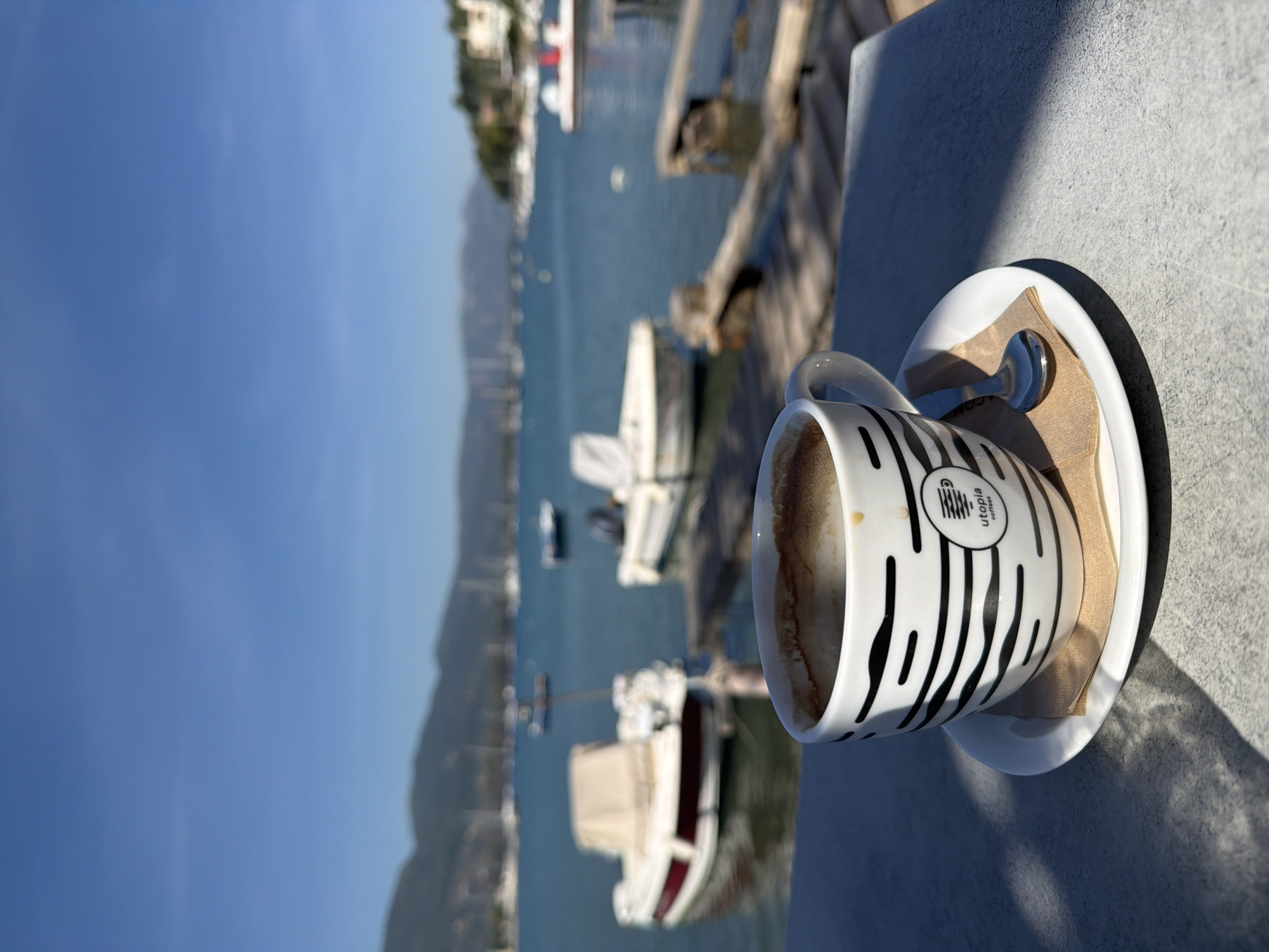 The image displays a serene outdoor scene, likely at a cafe or balcony overlooking a bay. In the immediate foreground, on a light grey, textured table, rests a white coffee mug adorned with distinctive black, zebra-like stripes. The mug, which appears partially consumed, is placed on a matching white saucer. A small silver spoon and a folded brown paper napkin are also visible on the saucer. The side of the mug features the text "utopia coffee" along with a logo.
Beyond the table, a vast, clear blue sky dominates the background. The middle ground features calm, deep blue water, characteristic of a sea or bay, dotted with several white boats and yachts, some docked and others moored. In the distance, hazy mountains line the horizon, with faint hints of buildings along the shoreline.
Bright sunlight casts distinct, long shadows from the mug and saucer onto the table, indicating a sunny day, likely during the morning or late afternoon. The overall ambiance suggests a peaceful moment of relaxation by the water, in a setting consistent with the provided context of Poros, Greece. The image is presented on its side, rotated 90 degrees clockwise from its natural horizontal orientation.