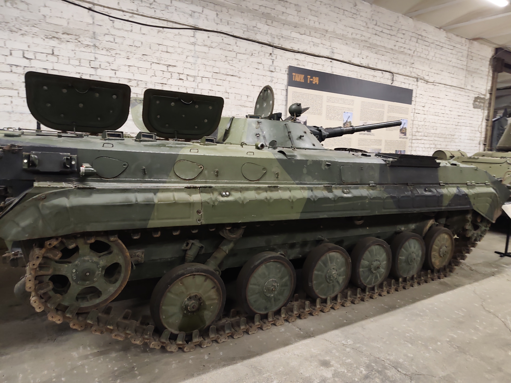The image shows the side view of a Soviet-era BTR-60 armored personnel carrier parked indoors. The vehicle is painted in a camouflage pattern of green and black. It has several road wheels and a continuous track system. Two hatches are open on the top of the vehicle, and a turret with a gun is visible. In the background, a white brick wall with some exposed brickwork is present, along with a sign that reads "TANK T-34". The floor appears to be concrete. This scene is located in the Viimsi Open Air Museum in Viimsi, Estonia, which displays military equipment. There are no people visible in the image.