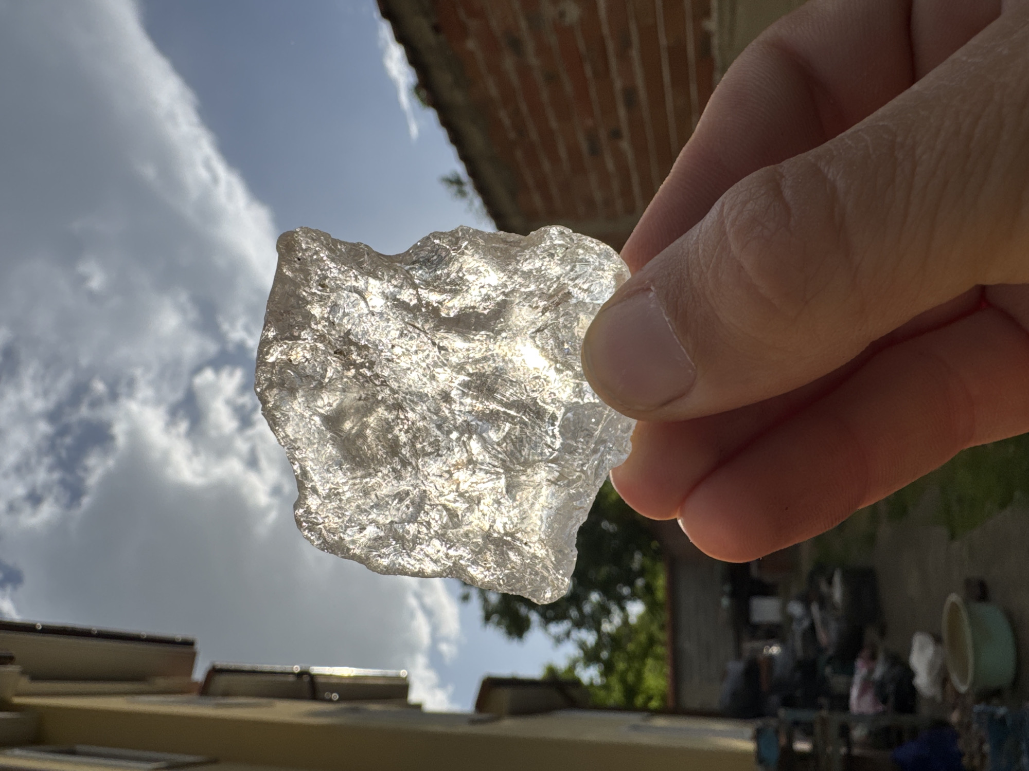 A person holds a translucent, irregularly shaped piece of what appears to be glass or ice in their right hand, with the thumb and forefinger visible. The background is an outdoor scene with partly cloudy skies. Below the hand and the object, the tops of buildings are visible, suggesting an urban environment. Other elements of the background include sections of brick and glimpses of trees. The location is Unknown, Unknown.