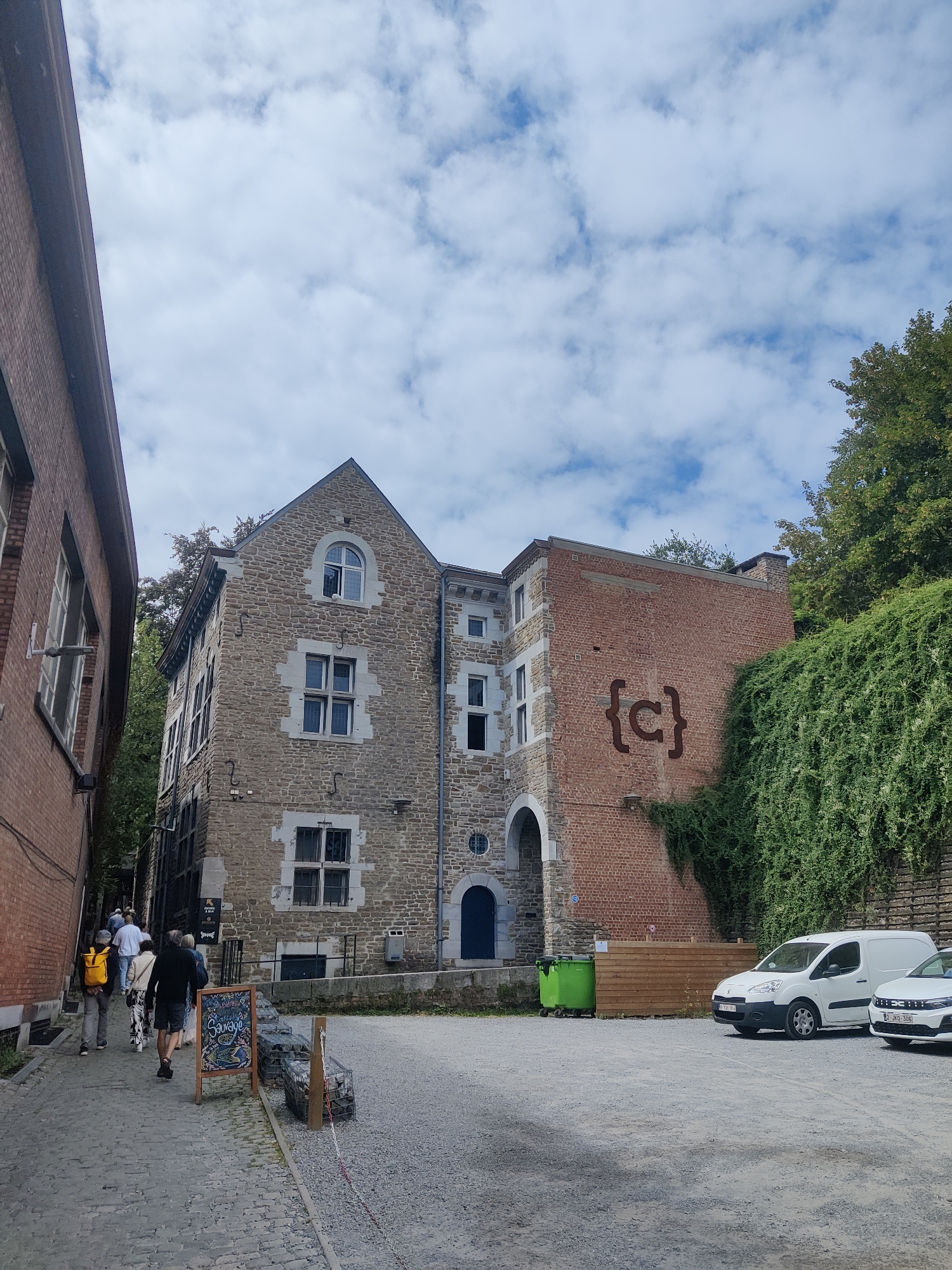 In Liège, Belgium, on a partly cloudy day, a group of people walks along a cobblestone street lined with brick and stone buildings. A brick building is on the left, and a multi-story stone building with a pointed roof is in the center, featuring multiple windows and decorative stone framing. To the right of the stone building is a red brick structure with a large, stylized "{c}" symbol on its facade. A white van and a white car are parked in a gravel lot to the right of the buildings. Lush green ivy covers a wall behind the brick building. A green trash bin is situated near the entrance of the stone building. A wooden sign with an illustration and text is placed near the cobblestone path.