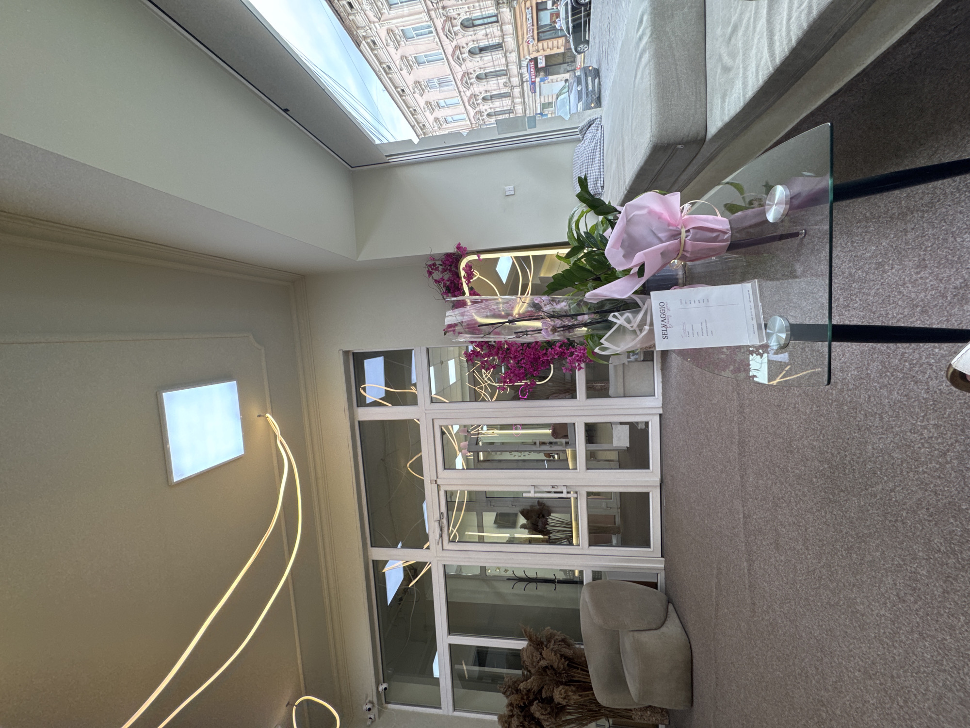 The image depicts the interior of a modern salon or boutique, likely located in Chernivtsi, Ukraine, based on the architecture visible through the large window. The space features a minimalist design with white walls and ceiling. Abstract, illuminated neon-like light fixtures are prominently displayed, curving across the ceiling and along the back wall.
In the foreground, a glass-topped coffee table with black metal legs sits on a textured brown carpet. On the table, a menu or price list for "SEL VAGGIO Beauty" is visible, along with a small bouquet of flowers wrapped in pink paper.
To the left, a beige, rounded armchair is positioned near a set of large glass doors, which appear to be the entrance to the establishment. Dried reeds in a tall vase are placed next to the armchair.
Through the glass doors, the interior of the salon can be seen, with more neon lighting and decorative plants. To the right, a large window offers a view of a European-style street scene with ornate building facades and passing cars. A cushioned seating area, resembling a bench or window seat, runs along the inside of the window. The overall atmosphere suggests a chic and contemporary commercial space.