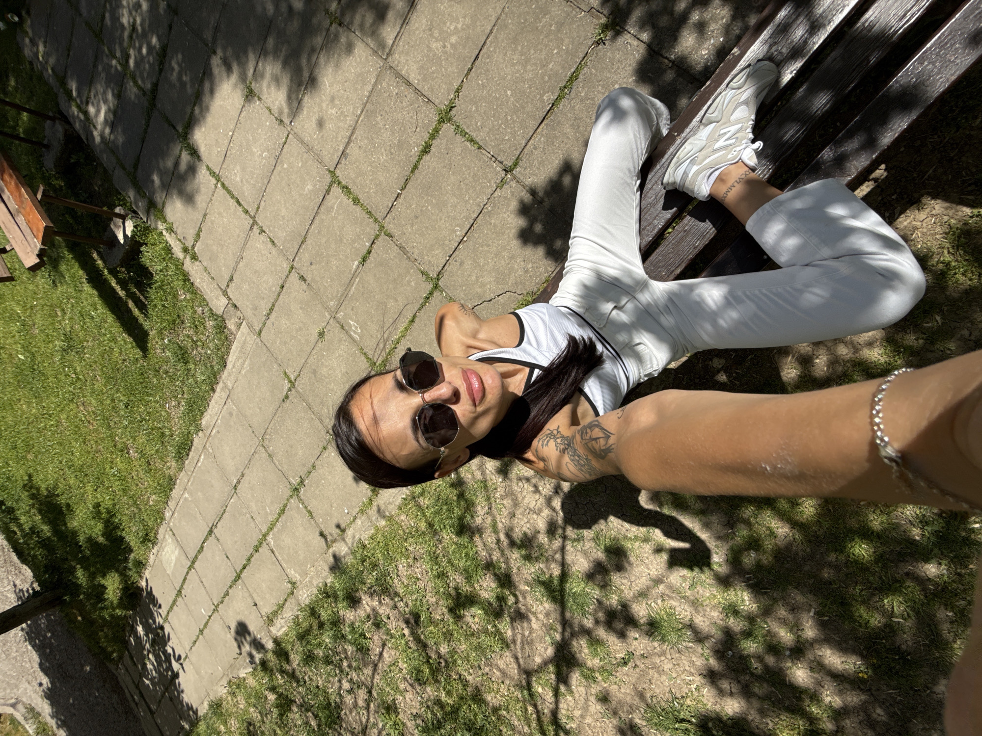 A woman is lying on a park bench in Pleven, Bulgaria. She is wearing white pants, a white top with black trim, white sneakers, and sunglasses. The bench is made of dark wood and is positioned on a paved pathway next to a grassy area. The scene is dappled with sunlight and shadows from nearby trees. The woman is holding her arm out, likely taking a selfie. A silver bracelet is visible on her wrist and there is a tattoo on her forearm. The background includes paving stones, grass, and parts of other benches or park furniture.