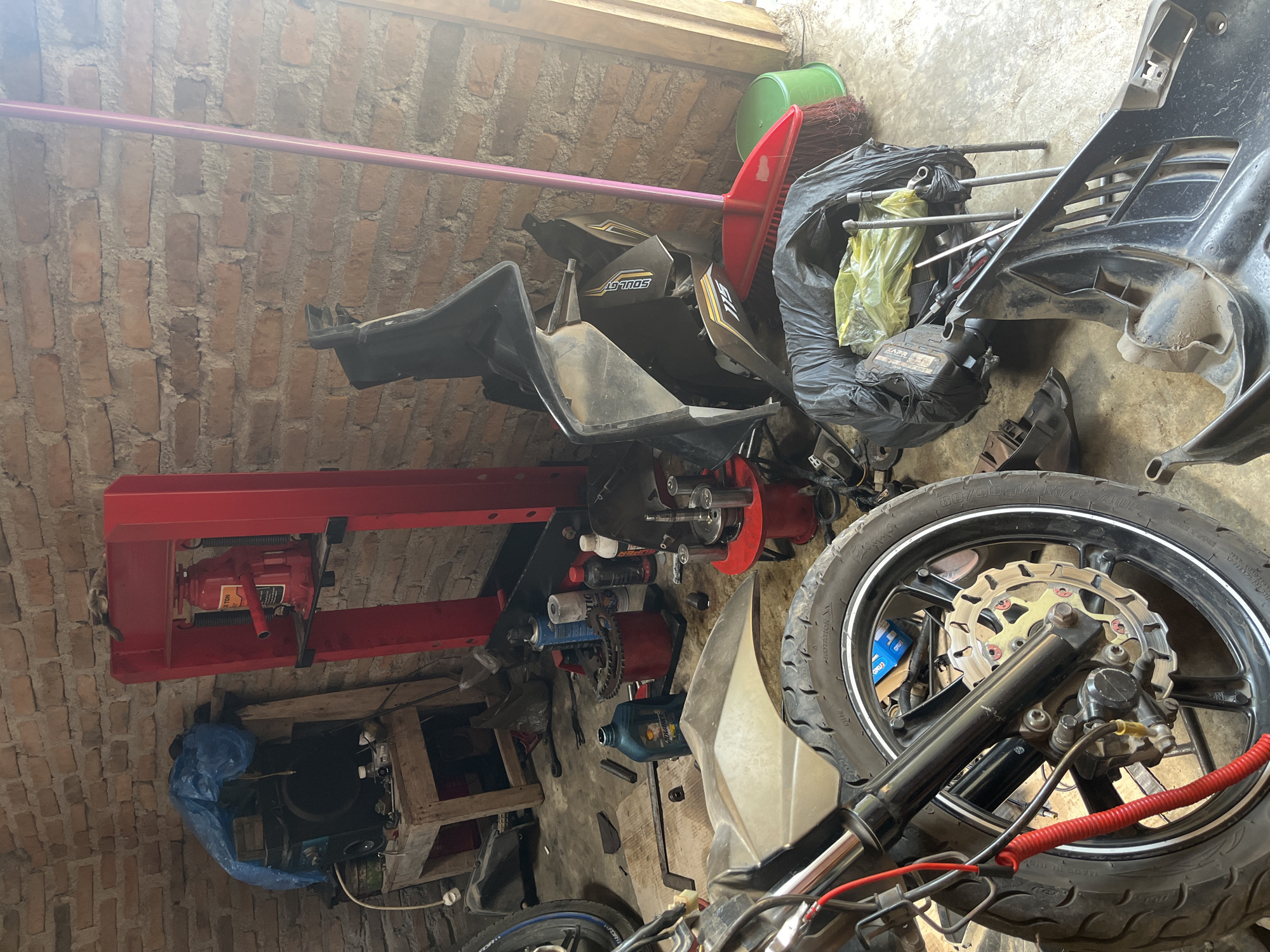 The image depicts a cluttered garage or workshop, likely located in Kebumen, Indonesia, with brick walls as the backdrop. In the foreground, a motorcycle wheel with a brake disc is visible. To the right, a red broom and a green bucket rest against the wall. Various motorcycle parts are scattered around the floor, including a large black plastic fairing and a black plastic bag filled with items, possibly tools and spare parts. A red hydraulic press with a jack is positioned against the wall. Other items visible include a bottle of oil, spray cans, and other mechanical components, suggesting ongoing motorcycle repair or maintenance. The lighting suggests it is daytime. There are no people visible in the image.
