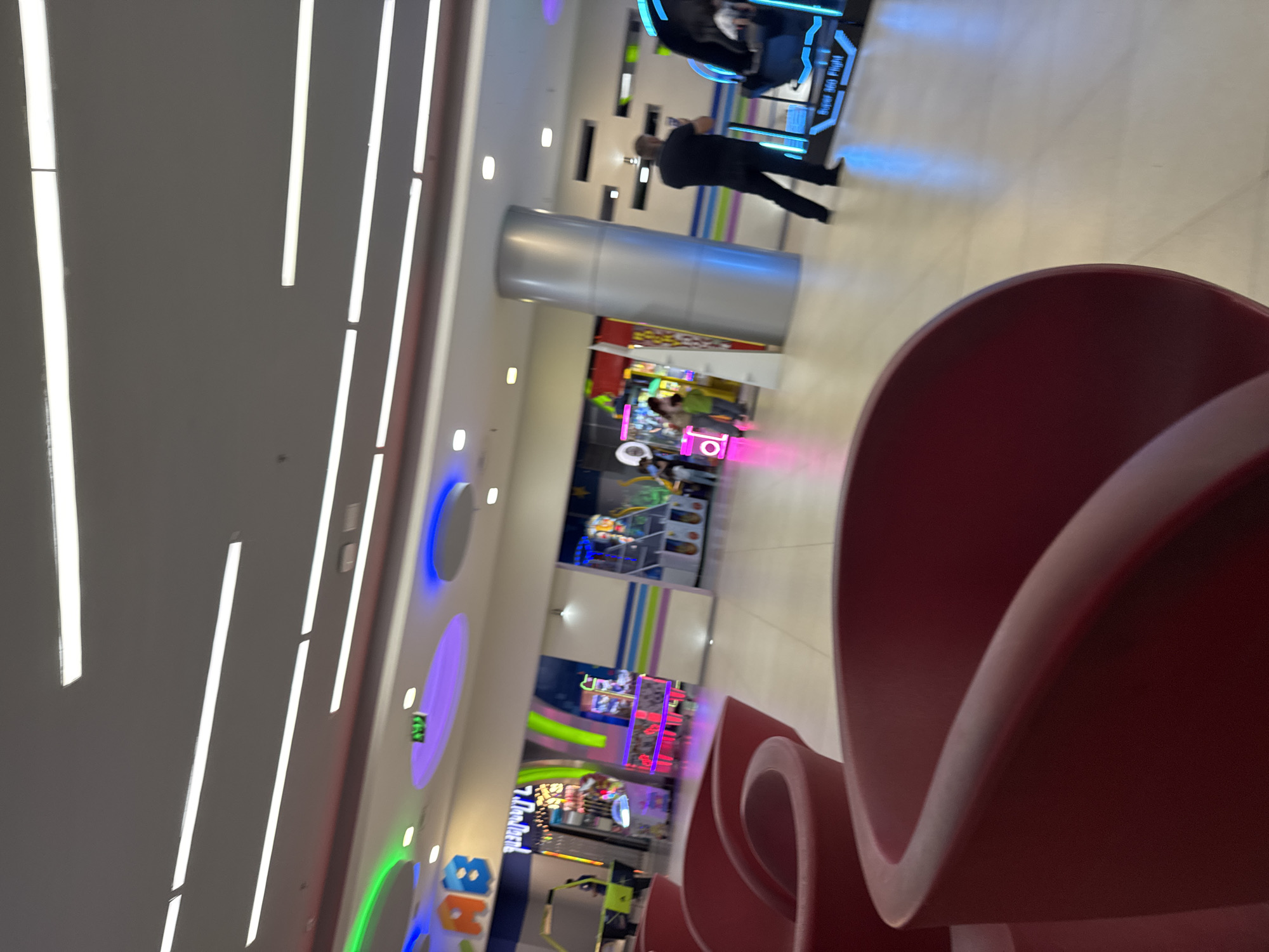 This image captures a vibrant, brightly lit indoor entertainment center, likely an arcade or amusement area within a mall, located in Yerevan, Armenia.
In the foreground, on the right, there is a large, sculptural piece of red seating or decoration, its smooth, curved surfaces partially visible. The mid-ground features a tall, silver cylindrical pillar. A man wearing a dark shirt and pants is seen walking past this pillar, with his back to the viewer. Behind him, other indistinct figures are visible further down the aisle.
The background is filled with various arcade games and amusement rides, characterized by their glowing, colorful lights in shades of blue, pink, and green. One game console clearly displays the text "Super Axis Flight." The walls are decorated with large, illuminated circular fixtures in blue, purple, and green, adding to the dynamic atmosphere. Part of a colorful sign with the letters "AB" is also visible in the lower left background.
The ceiling is adorned with numerous long, bright strip lights, providing ample artificial illumination. The overall impression is one of a modern, lively recreational space, with people engaging in or walking past the various activities.