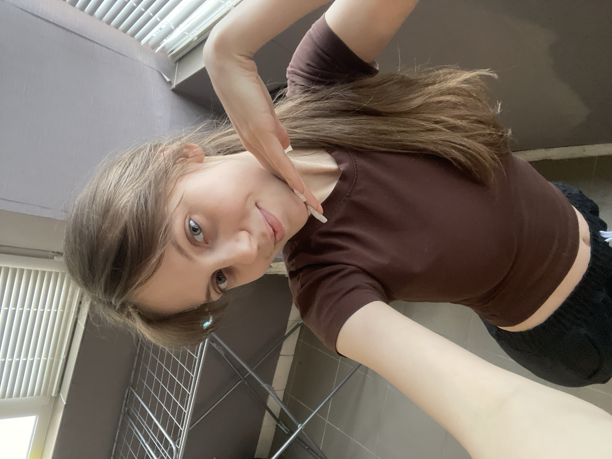 A young woman is taking a selfie indoors. She has light brown hair and fair skin, wearing a brown t-shirt and black bottoms. Her left hand is raised near her face. Behind her, there is a window with white blinds, and a metal drying rack stands against a gray-tiled wall. The scene appears to be in a room with gray tiles, possibly in Ufa, Russia, but there's no specific landmark or identifier to confirm the location definitively.