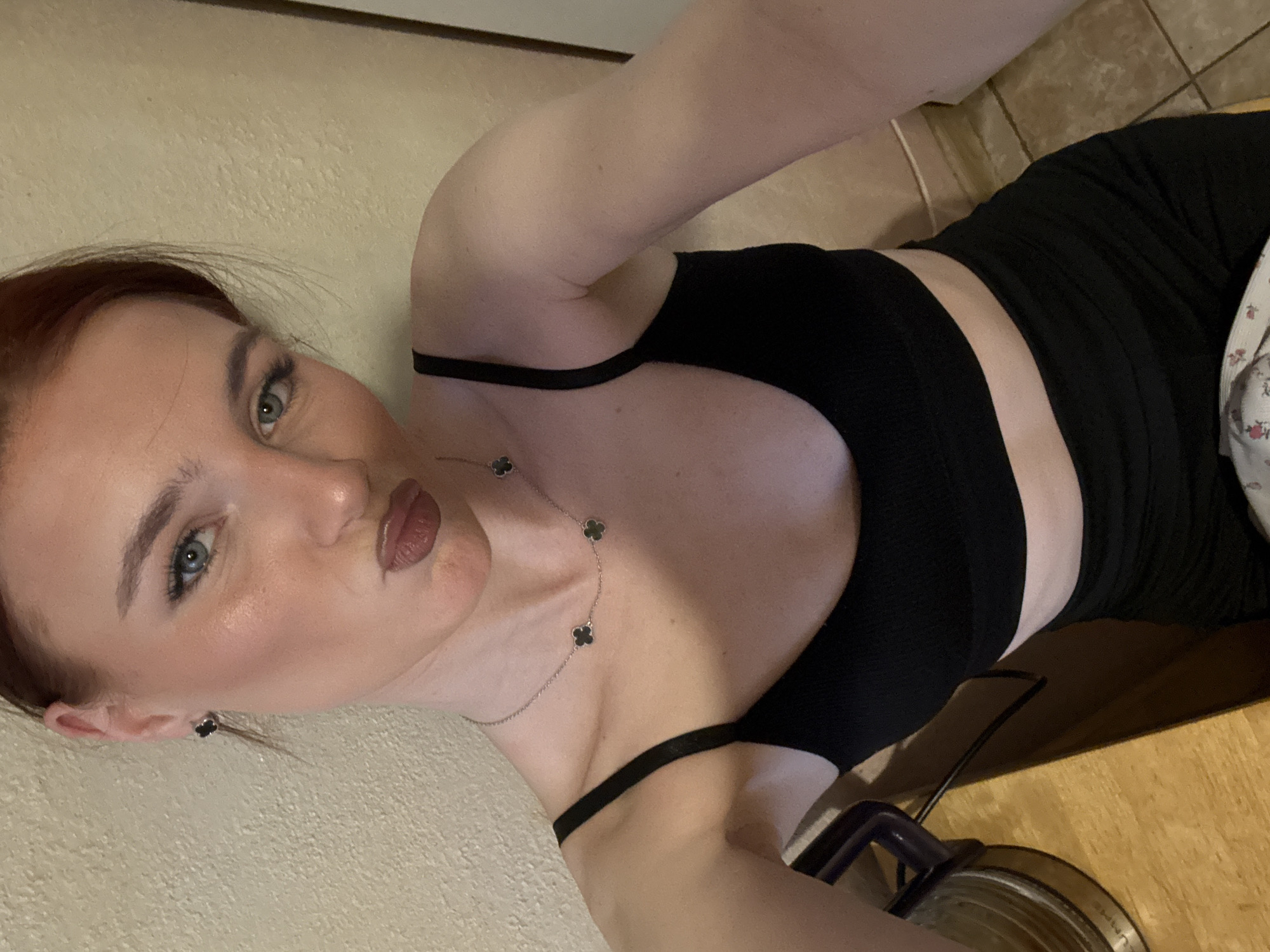 A fair-skinned woman with reddish hair is lying down, facing the camera. She is wearing a black tank top and black bottoms. She has on a necklace with clover-shaped pendants and matching earrings. The background consists of a light-colored wall and tiled floor. Part of a metal canister or appliance is visible near her body. The location is purported to be Murino, Russia.