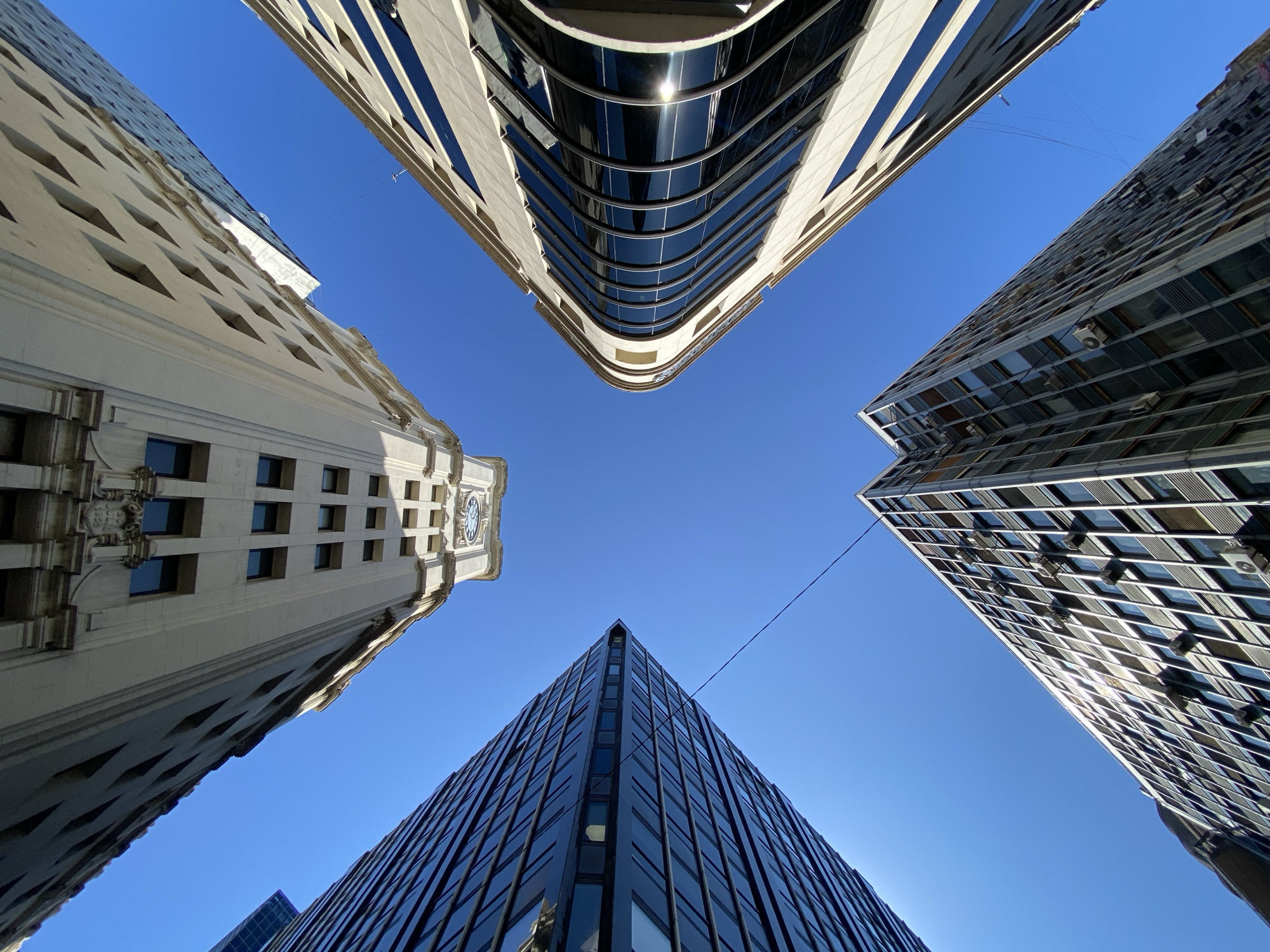 The image shows a worm's-eye view of four buildings in Buenos Aires, Argentina, with a clear blue sky above. The buildings are arranged in a square pattern around the central point of view. The building on the left is light-colored with a clock at the top. The building on the top is white with a curved facade. The building on the right is a dark-colored with many windows. The building on the bottom is the tallest and is dark colored with many windows. The buildings appear modern and well-maintained. There are wires strung between some of the buildings. The scene appears to be taken in daylight.