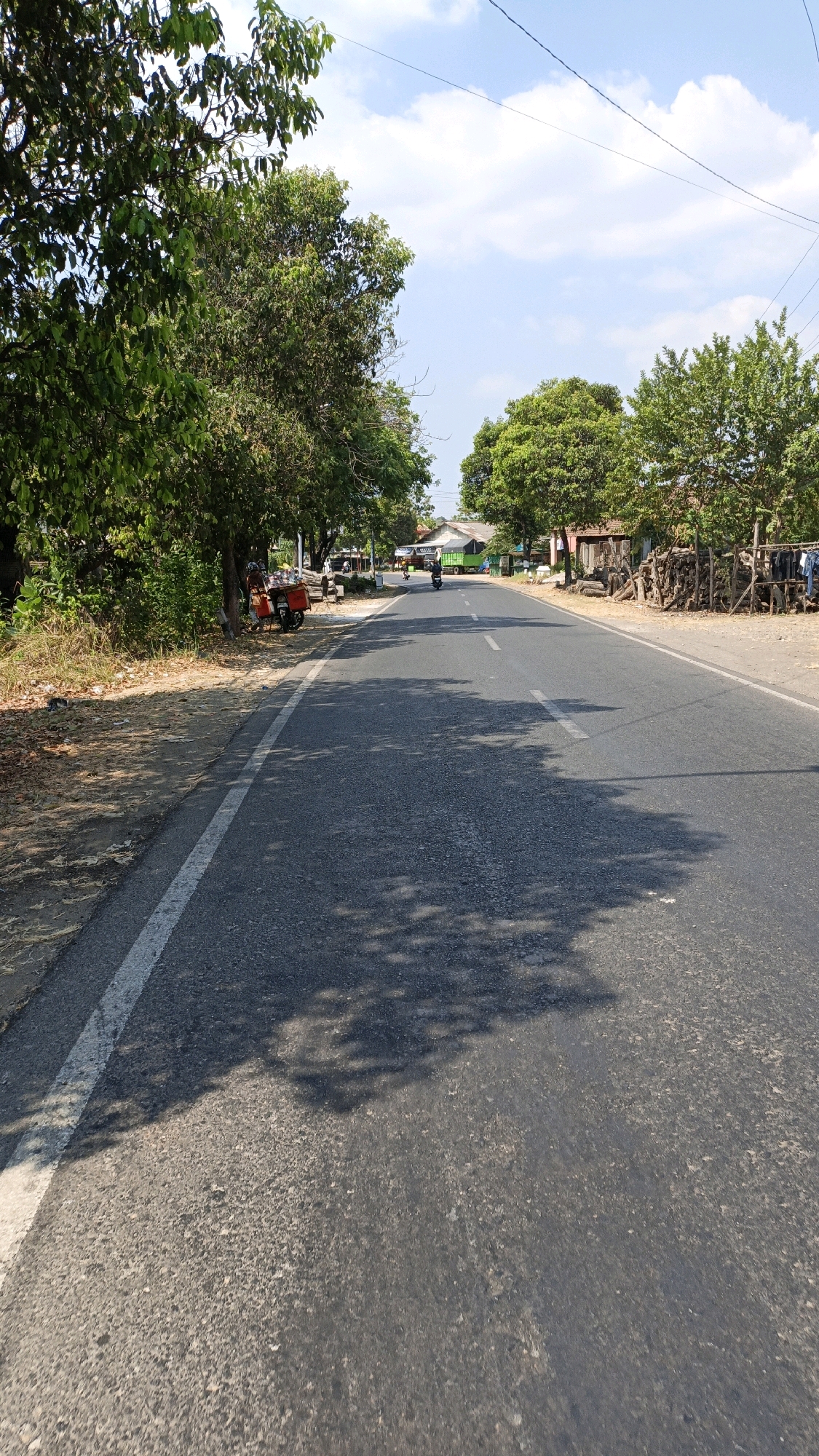 The image depicts a paved road in Sumbring, Indonesia, on a clear day with scattered clouds. On the left side of the road, there is a line of trees and foliage. A red motorcycle with a sidecar, possibly a mobile vendor, is parked by the side of the road beneath a tree, with a person visible sitting on it. Further down the road, more trees line the right side, with some structures and a pile of firewood. Vehicles are visible in the distance, including a truck and motorcycles. Shadows of trees are cast across the asphalt road, indicating the presence of sunlight filtering through the leaves. The road has a white line marking the edge on the left and a broken white line down the center. The overall scene suggests a rural or semi-rural setting along a main thoroughfare.