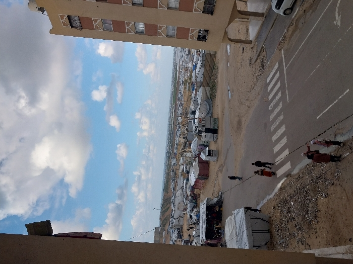 This elevated wide shot captures a street scene bordered by temporary shelters and permanent buildings in Khan Yunis, Palestinian Territory, under a partly cloudy blue sky.
In the foreground, the edge of a light-colored building is visible on the left, with a red object protruding. On the right, a multi-story, light-brown building with decorative geometric patterns on its facade rises, featuring an open ground floor supported by arches. A white car is parked beneath this building on the right.
A paved road with a white-striped pedestrian crossing (zebra crossing) runs horizontally through the midground. Three individuals are crossing the street on the zebra crossing from left to right: one individual in dark clothing, followed by an individual in a red garment and another in dark clothing. Further down the road, on the opposite side, another individual in dark clothing walks near the temporary structures. Two women stand on the dirt shoulder in the lower-left, facing towards the road and the distant structures; one wears a brown garment, the other a light headscarf and dark top.
To the left of the road, a large white tent-like structure is situated near smaller, makeshift stalls covered with various tarpaulins (black, white, red). Beyond these immediate structures, a sprawling camp of numerous, densely packed makeshift shelters and tents, predominantly grey and white, stretches across the sandy terrain into the distant background. The horizon reveals a flat landscape with indistinct distant structures beneath the scattered white clouds in the bright blue sky.