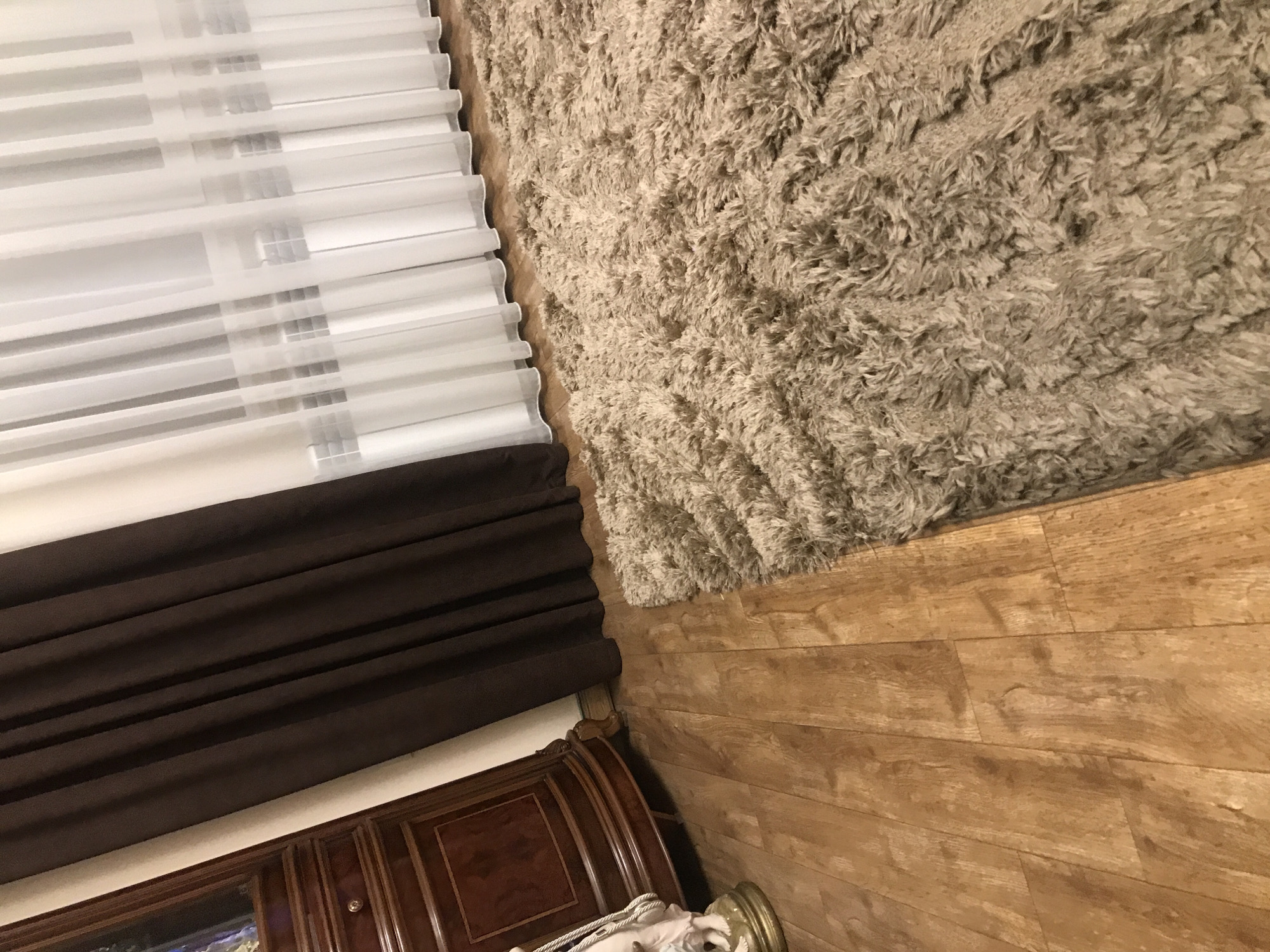 The image depicts an interior space. Dominating the upper-left is a window covered by horizontally-oriented, white blinds with brown drapery below. Adjoining the window blinds on the right is a shag rug in light brown, extending to the upper-right corner. The remaining space is filled by hardwood flooring. A piece of dark wooden furniture is visible in the lower-left corner. The environment is indoors. There is no evidence of a specific location in Yerevan, Armenia, or any details related to people, actions, or interactions.