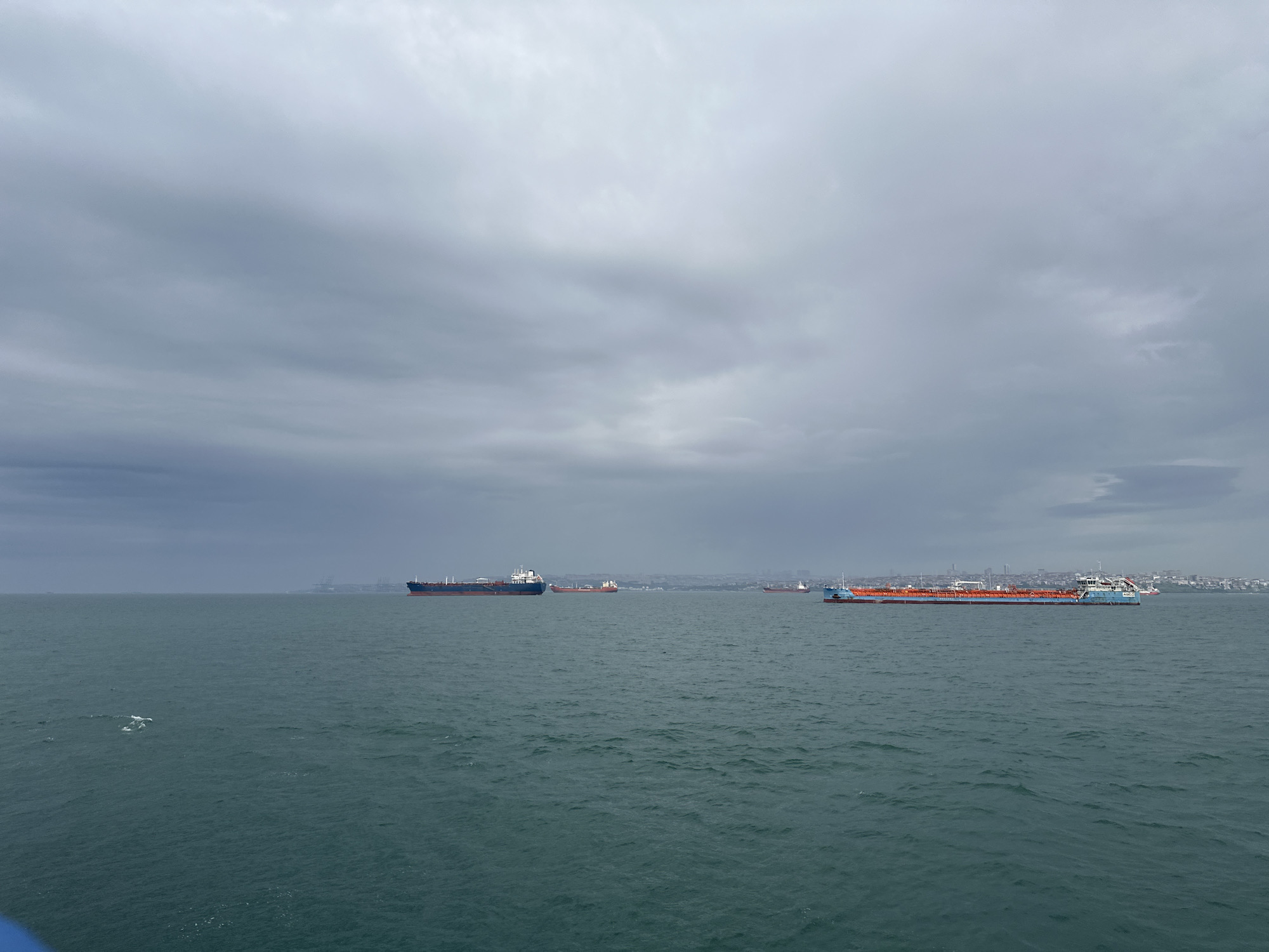 The image depicts a seascape under a cloudy sky in Unknown, Türkiye. Several ships are visible on the water. In the distance, a coastline with buildings is discernible. The sea is relatively calm, with small waves.