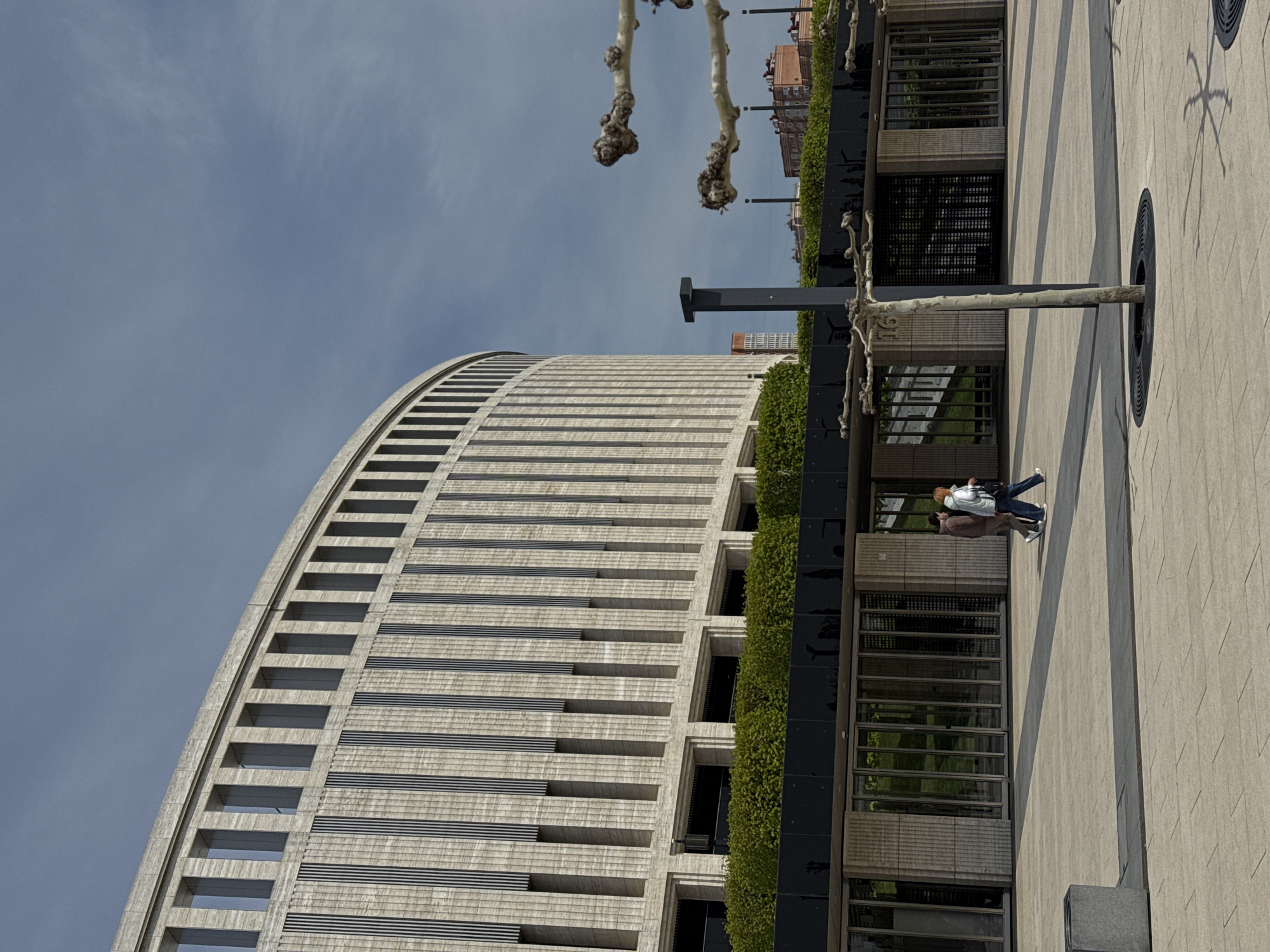 An outdoor, eye-level photograph depicts a modern urban scene dominated by architecture and a public plaza under a clear sky.
On the left, a large, multi-story building with a distinctive curved facade rises prominently. Its exterior is constructed from light-colored material, likely concrete or stone, featuring a series of prominent horizontal bands and recessed windows or openings that create a ribbed texture. Adjacent to this curved section, the building transitions to a flatter, vertical design characterized by dark, reflective glass panels and vibrant green vertical landscaping, resembling hedges or a green wall.
In the mid-ground and foreground, a wide, paved pedestrian area or plaza extends across the frame. The paving consists of light-colored square tiles arranged with darker, linear strips, forming a subtle geometric pattern. Several deciduous trees with bare, gnarled branches are spaced along this walkway, each emerging from a black, circular grate set into the pavement.
Two people are seen walking casually on the paved area. One individual wears a light-colored jacket or hoodie and dark pants, while the other is dressed in a brown jacket and dark pants. They appear to be walking side-by-side, moving towards the left side of the frame.
The sky is a bright, clear blue with a few very faint, wispy clouds. Strong sunlight illuminates the scene, casting distinct shadows from the trees and structures onto the paved ground, indicating a sunny day, likely mid-morning or afternoon. Other buildings are visible in the background, beyond the primary structures. There is no visible text in the image.