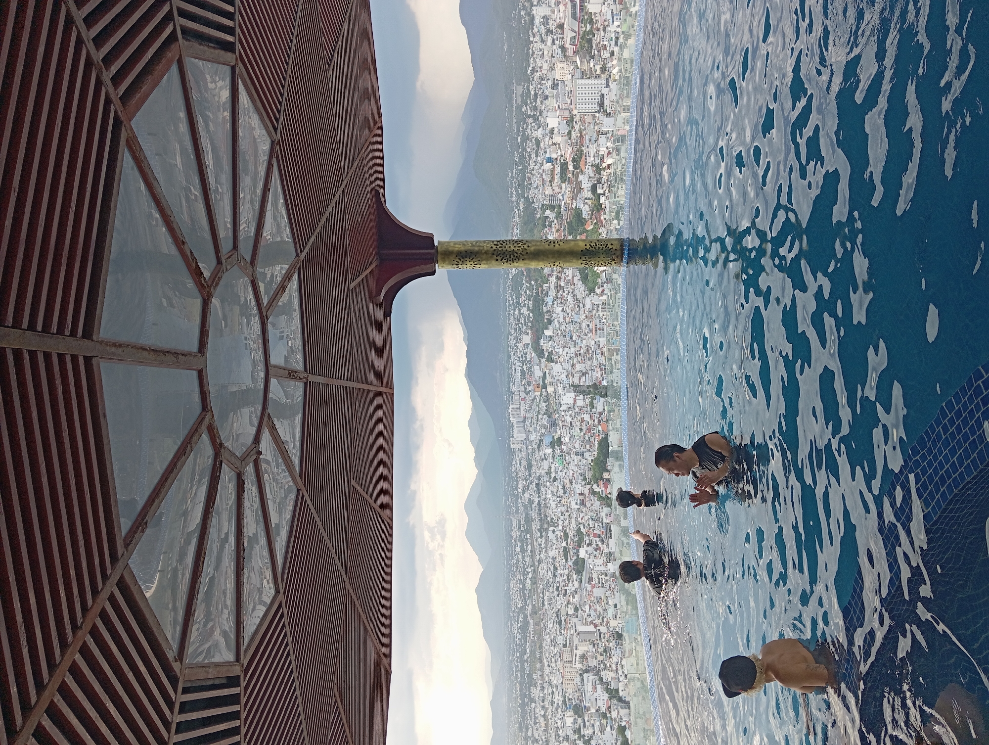The image depicts a rooftop infinity pool overlooking a dense cityscape with mountains in the background. Four individuals are visible in the pool. One person, wearing a black cap, is facing away from the camera on the left. To their right, another person is in the water, looking towards the city. In the center, a person in a black shirt is pointing towards the cityscape. On the right, a person with their hands clasped is looking towards the center. The pool features blue tiled edges and is surrounded by a modern architectural structure with a wooden ceiling and a decorative column. The city appears to be Nha Trang, Vietnam, based on the architectural style and the mountain range typical of the region. The sky is overcast, suggesting potential rain or dramatic weather.