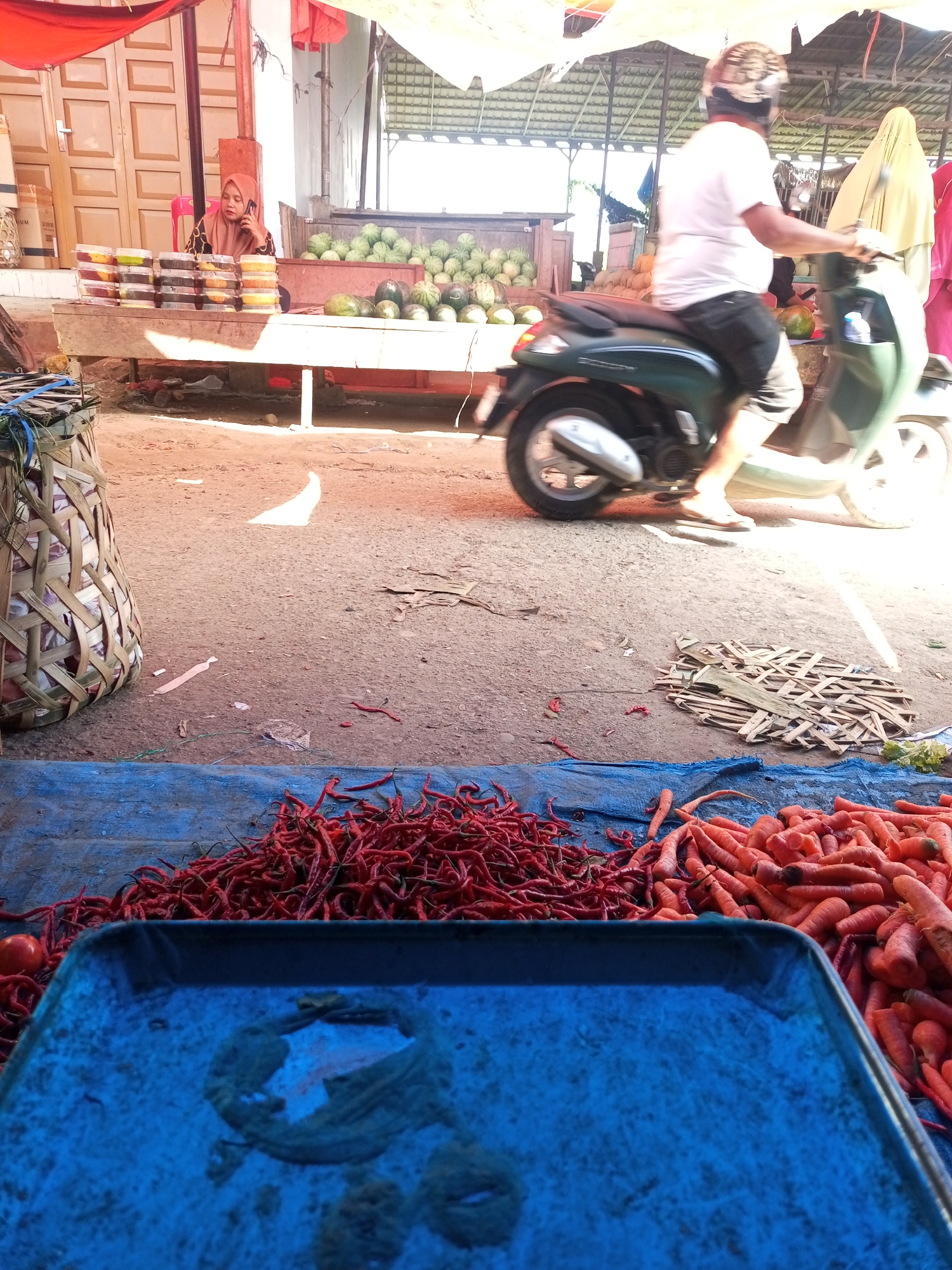The image depicts a street market scene in Lhokseumawe, Indonesia, likely on a sunny day given the strong shadows. In the foreground, a blue tray sits in front of a display of fresh red chilies and carrots laid out on a blue tarp. To the left, a large woven basket filled with items, possibly onions, is visible. In the mid-ground, a person wearing a white t-shirt and black shorts, riding a dark green scooter, passes through the market. The scooter's passenger, wearing a helmet with a patterned design, is slightly blurred due to motion. Behind the scooter, a person in a yellow hijab is seen. In the background, a woman wearing a peach-colored hijab is seated and talking on a mobile phone, with stacks of food containers on a wooden table in front of her. Behind her, a large pile of watermelons is displayed on a similar table. The market stalls are covered with tarps, and the ground is a dirt or gravel surface. The overall impression is that of a bustling local market with a variety of produce and activity.