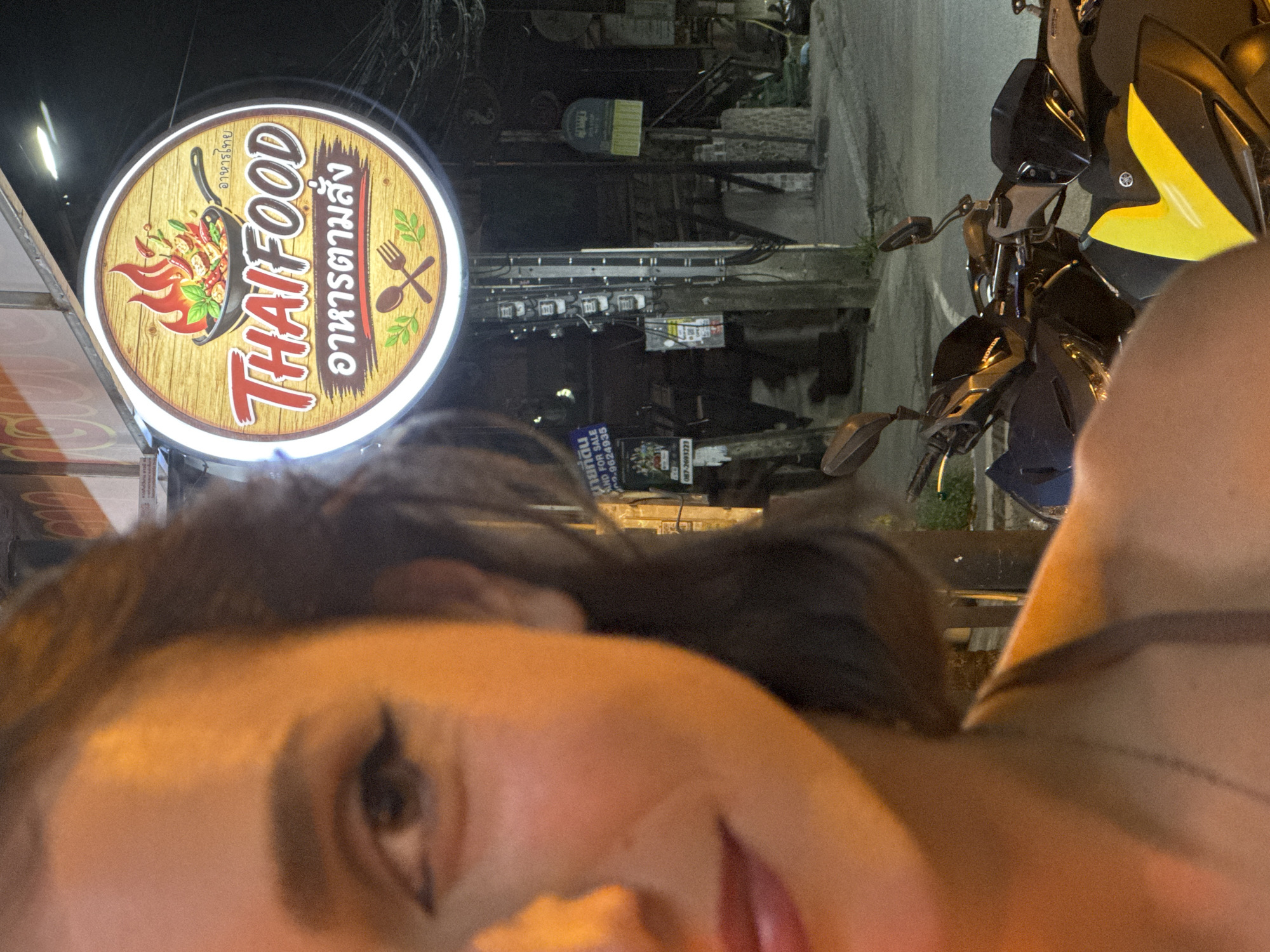 The image shows a close-up of a woman's face on the left side, with the right side of the image showing a brightly lit sign for "THAI FOOD" with a wok full of food and flames. The sign is circular with a wooden texture and a neon border. Below the food illustration, Thai script is visible. Further down the sign, a spoon and fork are depicted. The sign is mounted on a pole with various electrical meters and wires. In the background, a street scene is visible at night, with a motorcycle in the foreground. The overall setting appears to be a street in Patong, Thailand, indicated by the signage and common streetscape elements. The woman's eye is visible with winged eyeliner, and her lips are slightly parted. The lighting suggests it's nighttime, with the sign being the primary light source in the immediate vicinity.
