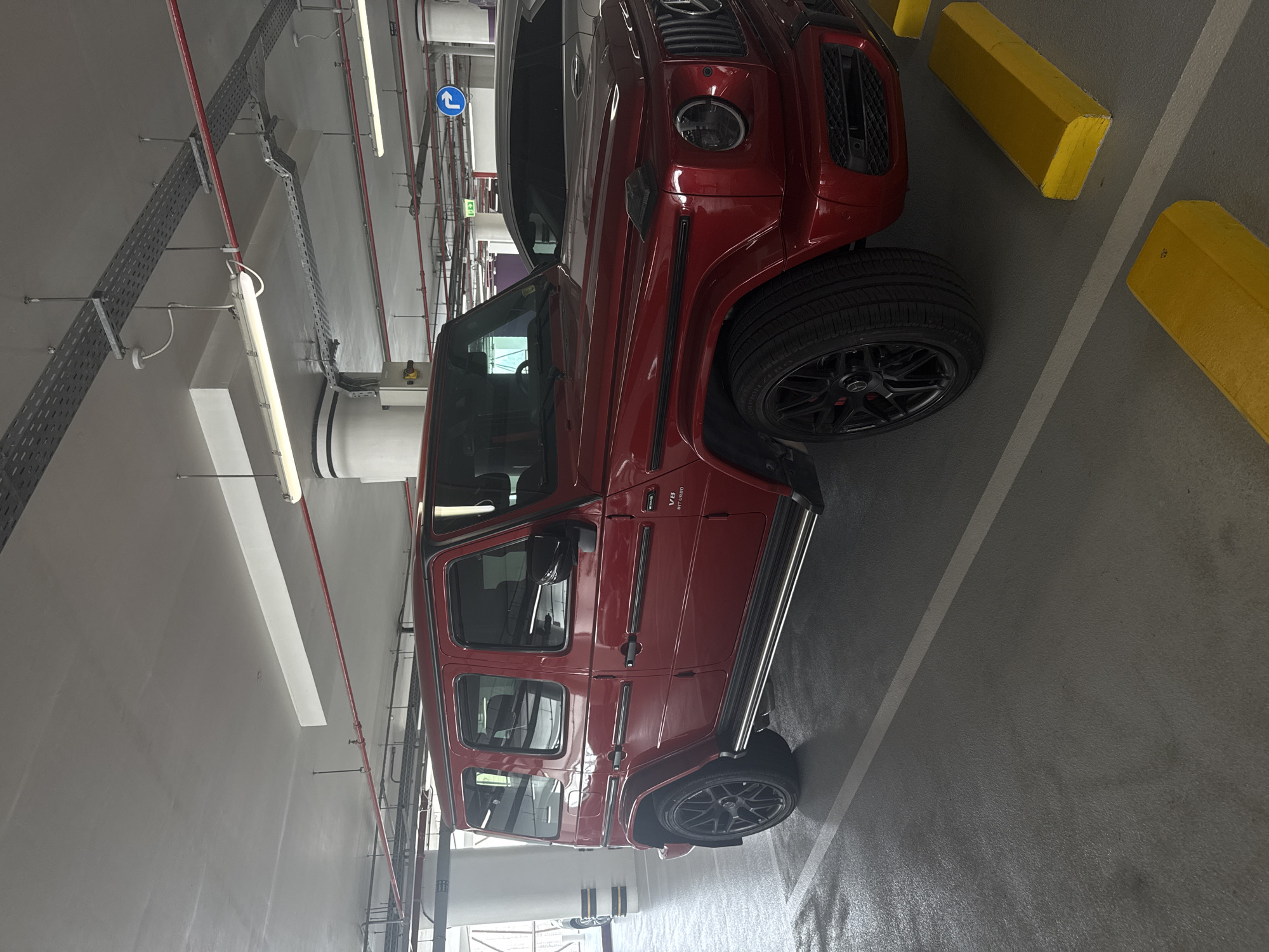 A glossy red Mercedes-Benz G-Wagen (G-Class) SUV is prominently parked in a modern, well-lit parking garage. The vehicle, likely a G 63 AMG, is visible from its front-right side, extending towards the rear. Notable features of the car include its distinctive grille with the Mercedes-Benz emblem, circular headlights, black multi-spoke wheels, and silver side steps. The side fender clearly displays "V8 BiTurbo" and "AMG" badging.
The parking environment consists of a clean, light grey concrete floor marked with white parking lines and yellow concrete parking barriers. The ceiling structure is visible, featuring exposed red pipes (likely fire suppression), silver perforated metal cable trays, and long fluorescent light fixtures providing artificial illumination. A blue circular sign with a white arrow pointing left is mounted on a column or wall in the background, indicating traffic flow. The overall setting appears to be a well-maintained, possibly upscale, indoor parking facility, consistent with locations in Dubai, United Arab Emirates. No people or specific activities are depicted; the scene captures a stationary vehicle in its parking spot.