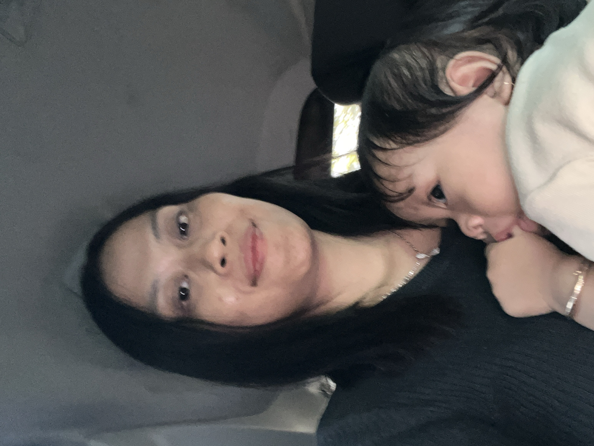 An over-the-shoulder selfie captures a woman with long, dark hair holding a baby in what appears to be the interior of a car. The woman's face is in the foreground, and she is smiling. The baby, positioned in front of her, is looking to the left and has its hand near its mouth. The environment is dimly lit and appears to be inside a vehicle, with car interior elements visible in the background. There are no identifiable landmarks or specific indicators of the city of Getas, Indonesia.