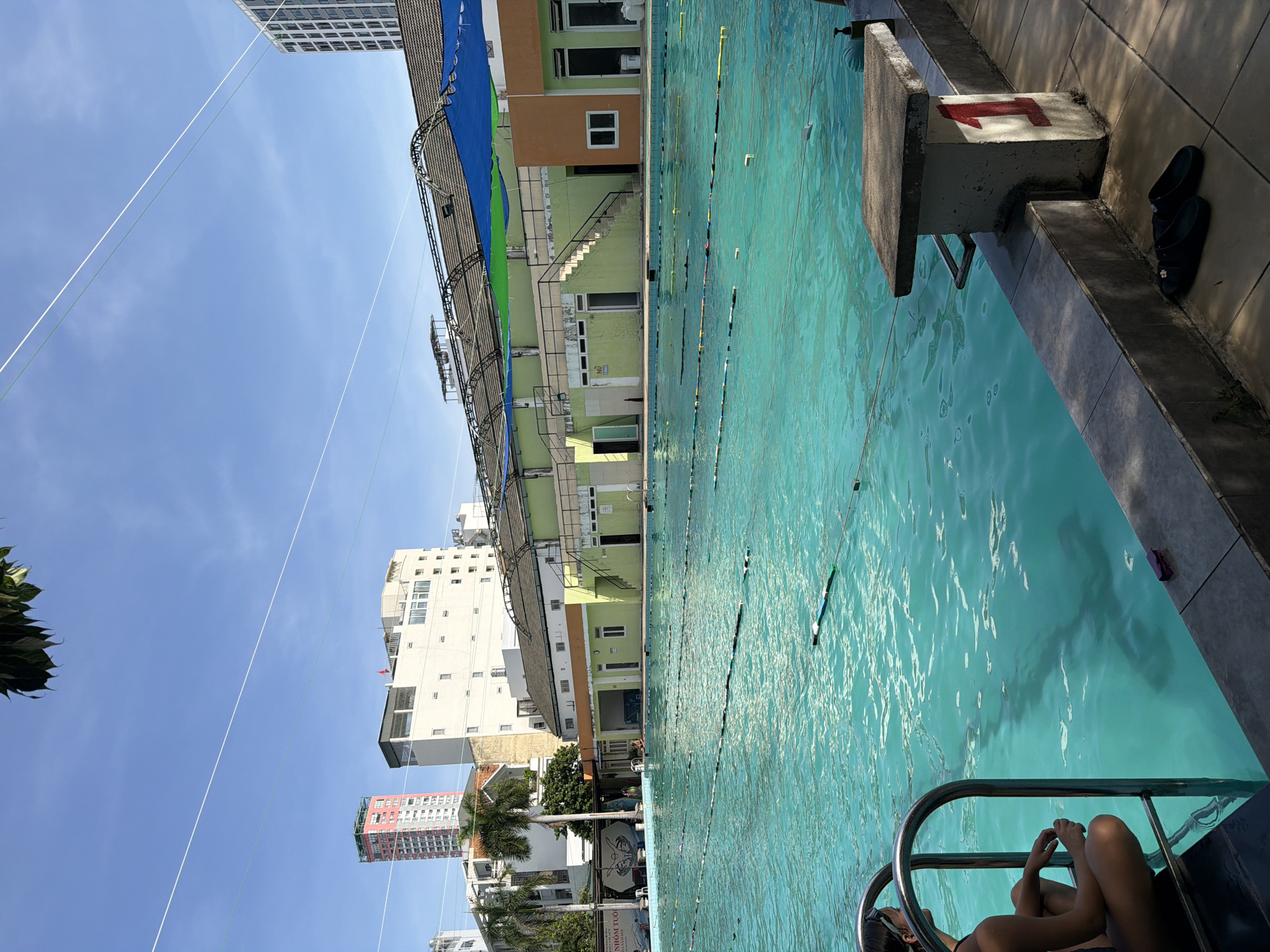 The image depicts a large outdoor swimming pool facility under a clear blue sky in Nha Trang Ward, Vietnam. The pool itself is filled with turquoise water, marked with lane lines and buoys. In the foreground, a woman with dark hair wearing a dark swimsuit is sitting on the edge of the pool, near a metal ladder. Her body is positioned in profile, facing towards the left side of the frame.
Adjacent to the pool's edge, a concrete diving block is visible, marked with a large red number "1" on its side. A pair of black flip-flops rests on the tiled ground next to the diving block. The background features a low-rise building with a green and orange facade, featuring numerous windows and doors. Stairs lead up to an upper level. Further in the distance, several tall buildings and palm trees are visible, indicating an urban setting. A large canopy structure with blue and green fabric is erected over part of the pool area. The overall scene suggests a public or recreational swimming facility on a sunny day.