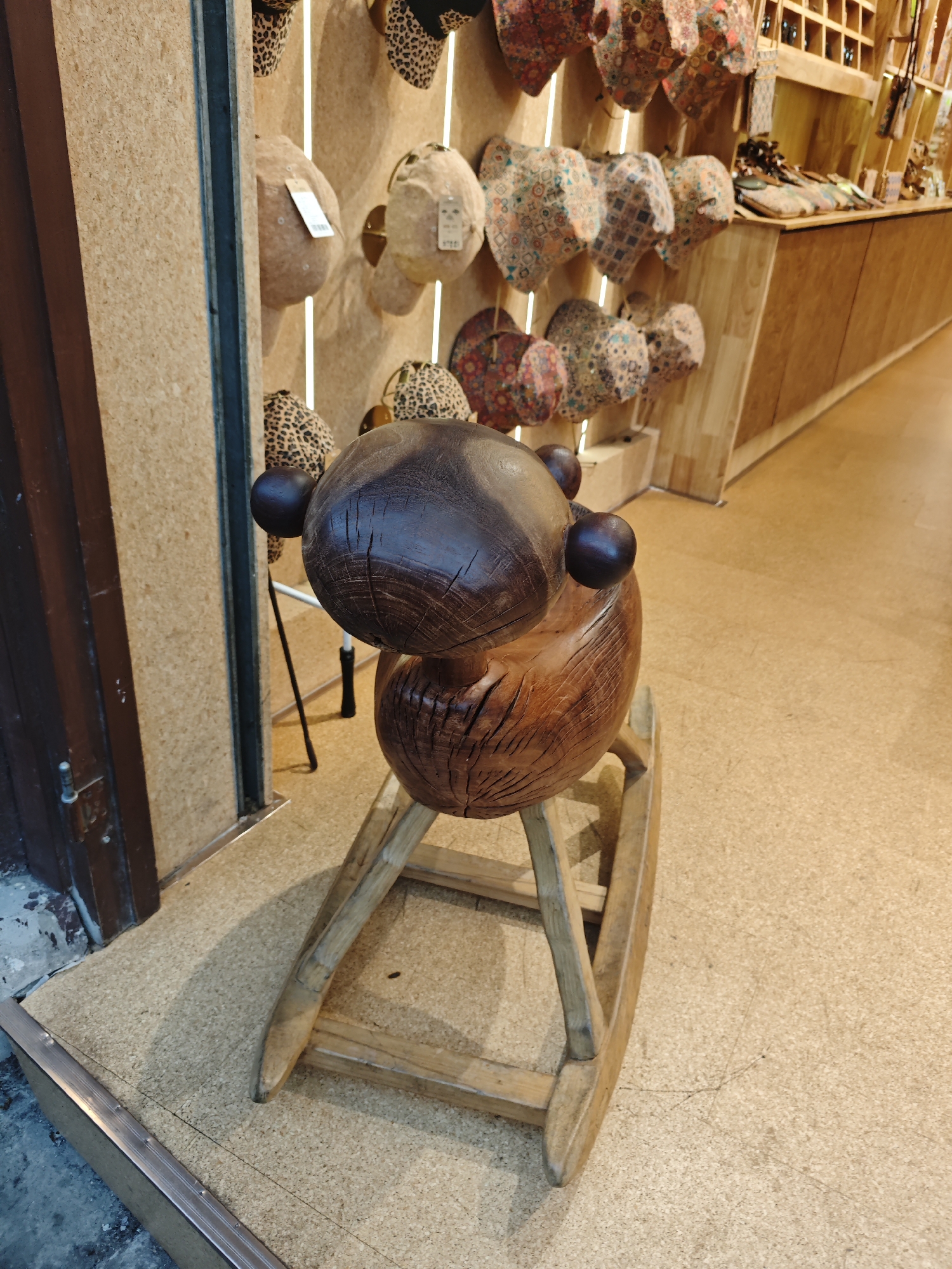 A high-angle, indoor shot depicts a wooden rocking horse in a commercial setting. The horse is crafted from dark and light brown wood with a visible grain pattern. The head is rounded with two spherical "ears" on either side. The rocking base is made of lighter, unfinished wood.
The horse sits near a storefront window with a dark brown frame. Inside the store, various hats and bags are displayed on the walls. The floor is a light brown, speckled material, possibly linoleum or a similar flooring.
The location is consistent with a retail store in an urban area, possibly in the Shapingba District, China.