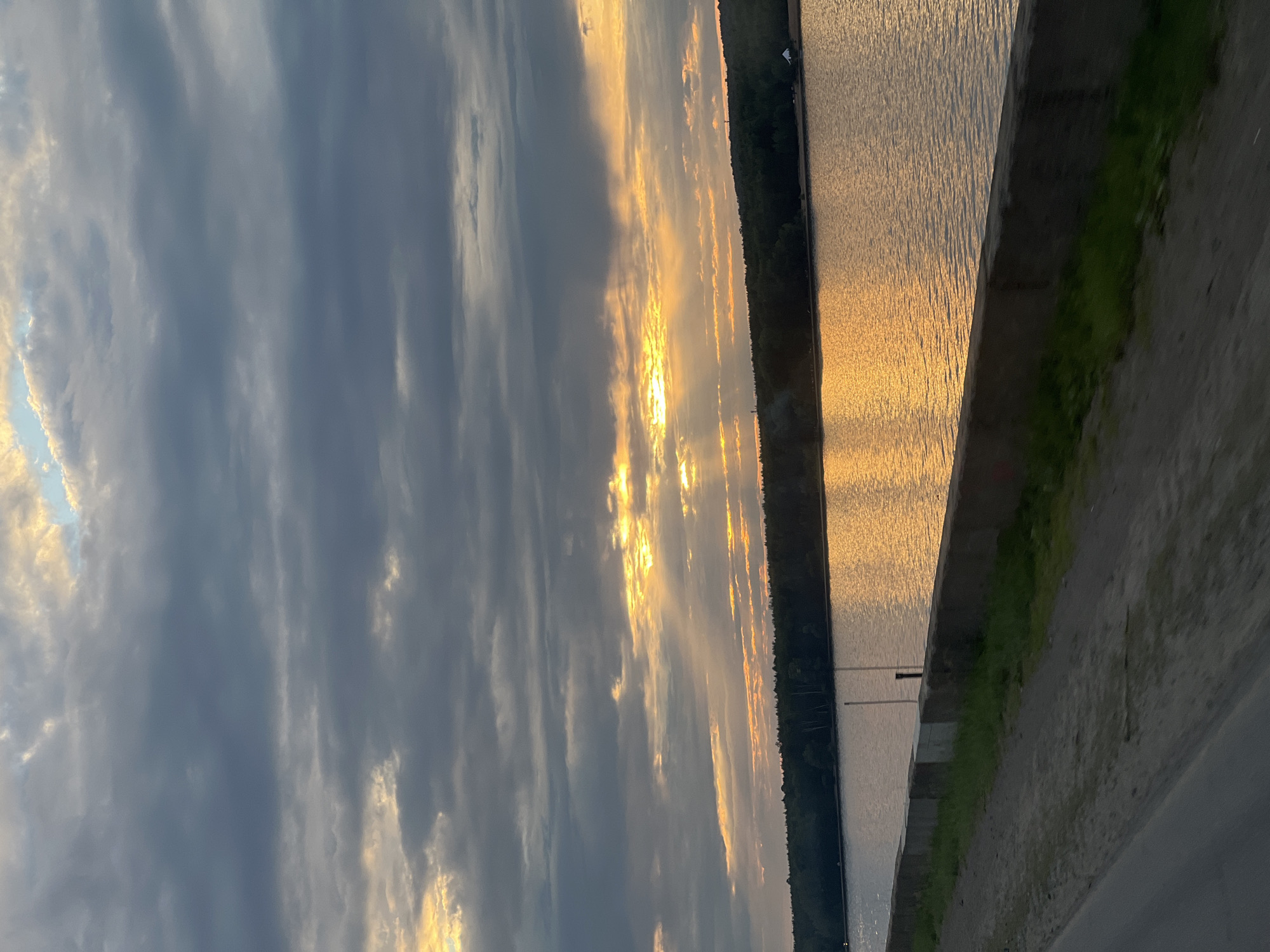 A sunset is captured over a wide body of water, reflecting golden light. Dark clouds dominate the sky, with rays of sunlight piercing through in some areas. A dense line of trees borders the far bank of the water. In the foreground, a concrete embankment separates a dirt path from the water. The image appears to be taken from a moving vehicle, as indicated by the blurred foreground. This scene likely depicts the Kama River in Chaikovsky, Russia, a common location for such views due to its expansive nature and surrounding forests.