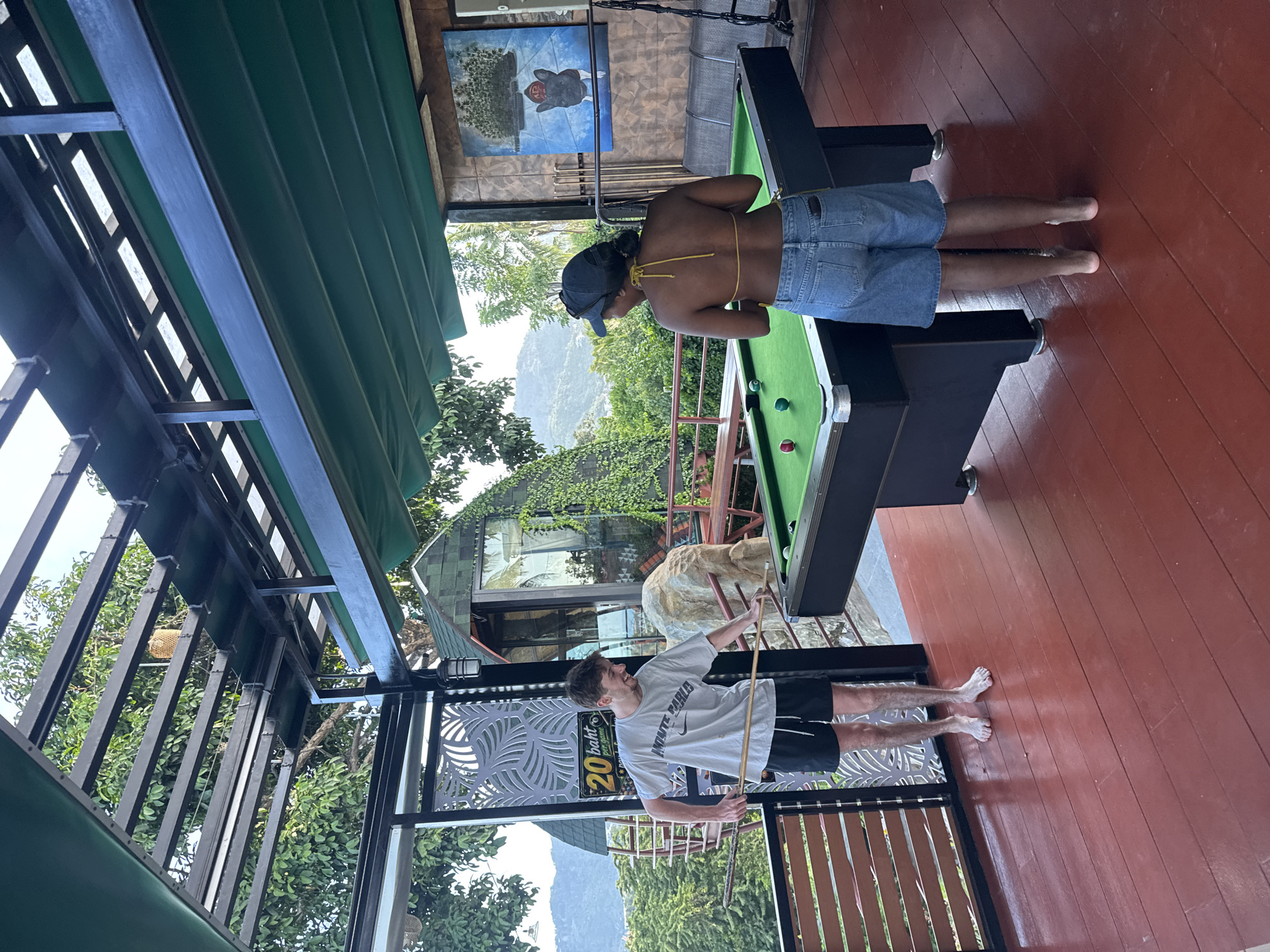 Two individuals, a male and a female, are playing billiards on an outdoor pool table. The male, wearing a white t-shirt and black shorts, stands on the left, holding a cue stick. The female, in a yellow bikini top and denim shorts, stands on the right. The pool table is green with several balls visible. The scene is on a wooden deck with a red floor, partially covered by a dark green awning supported by a metal frame. Lush green foliage and mountains are visible in the background, suggesting a tropical location. A small building with a green roof is also present. A sign displaying "20 baht per game" is mounted on a decorative panel to the left of the male. The environment is consistent with an outdoor recreational area in Ko Samui District, Thailand, likely a resort or guesthouse given the scenic backdrop.