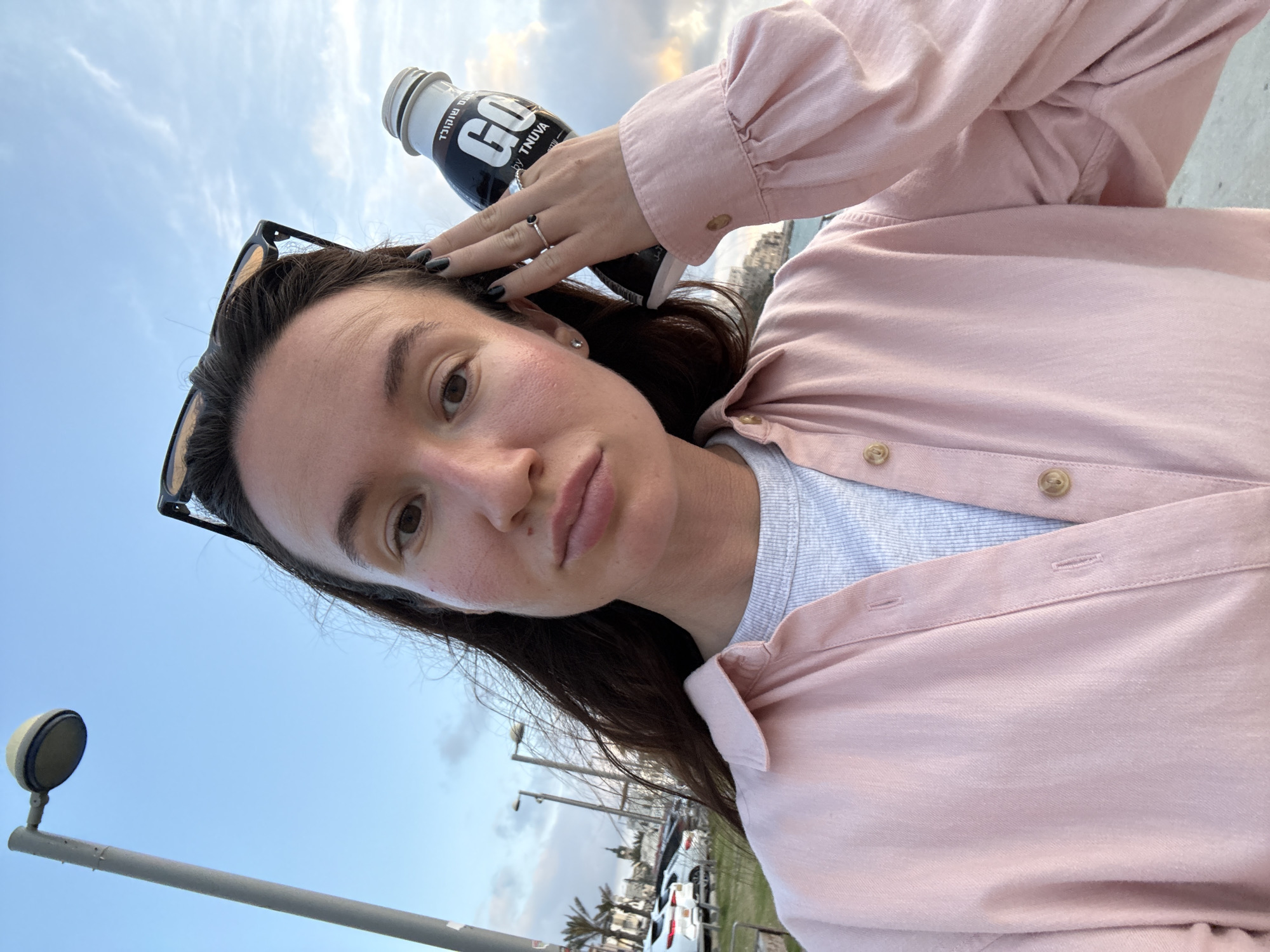 The image shows a person taking a selfie outdoors. The individual has fair skin, dark hair, and is wearing sunglasses on their head. They are wearing a pink button-down shirt over a light-colored shirt. A black bottle labeled "GO by TNUVA" rests on their head. The person is touching the bottle with their left hand, which has black nail polish and a ring. In the background, there are street lights, cars, palm trees, and buildings, suggesting a city location. Given the information, the location is Tel-Aviv, Israel. The image seems to be taken in a public space such as a promenade or park area, with the sea and sky visible in the distance.