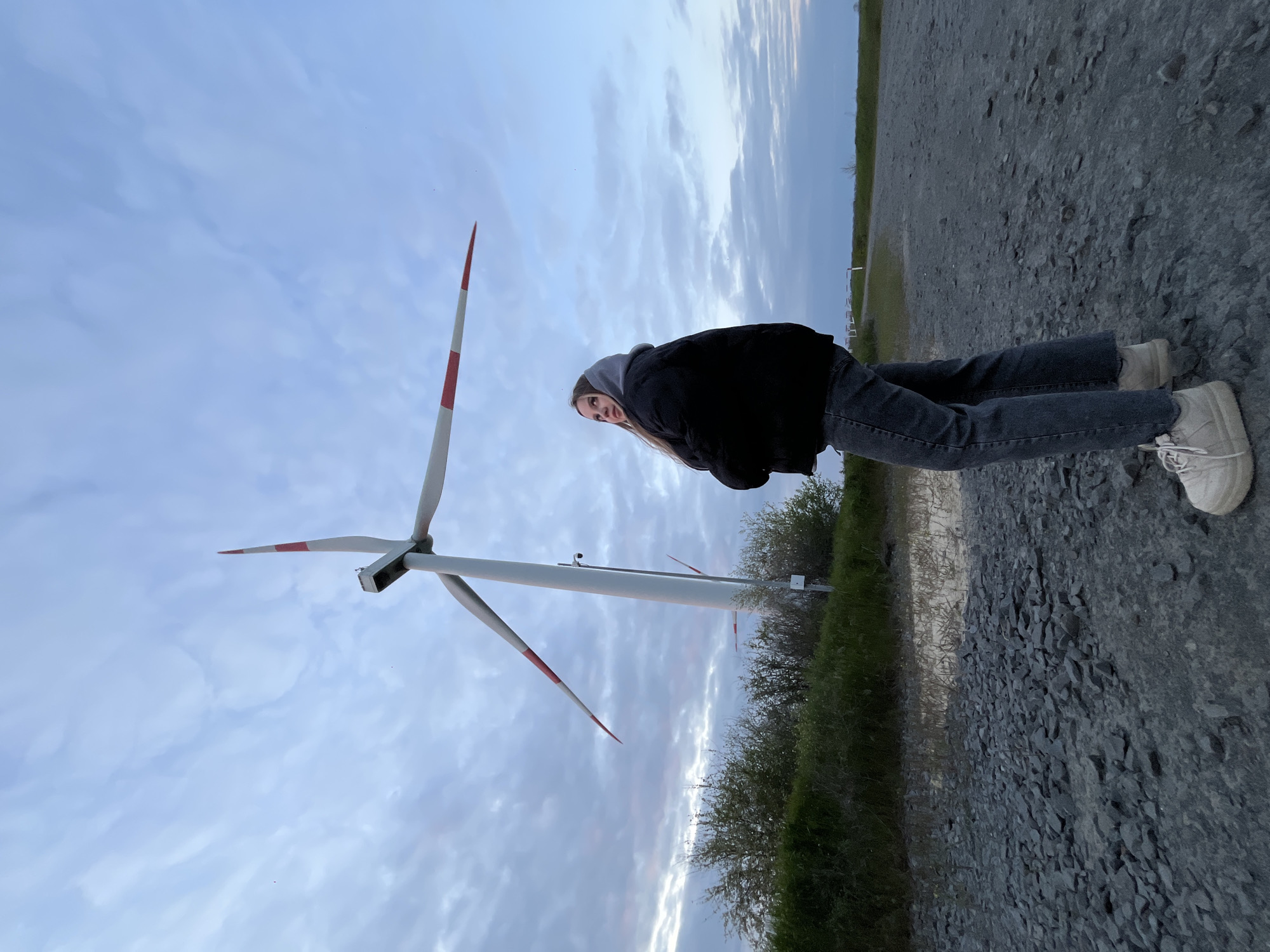 A young woman stands next to a wind turbine on a cloudy day. The woman wears a black jacket, gray jeans, and white shoes. The ground is covered in gravel. The wind turbine has three blades with red tips. The location is in Port-Katon, Russia.