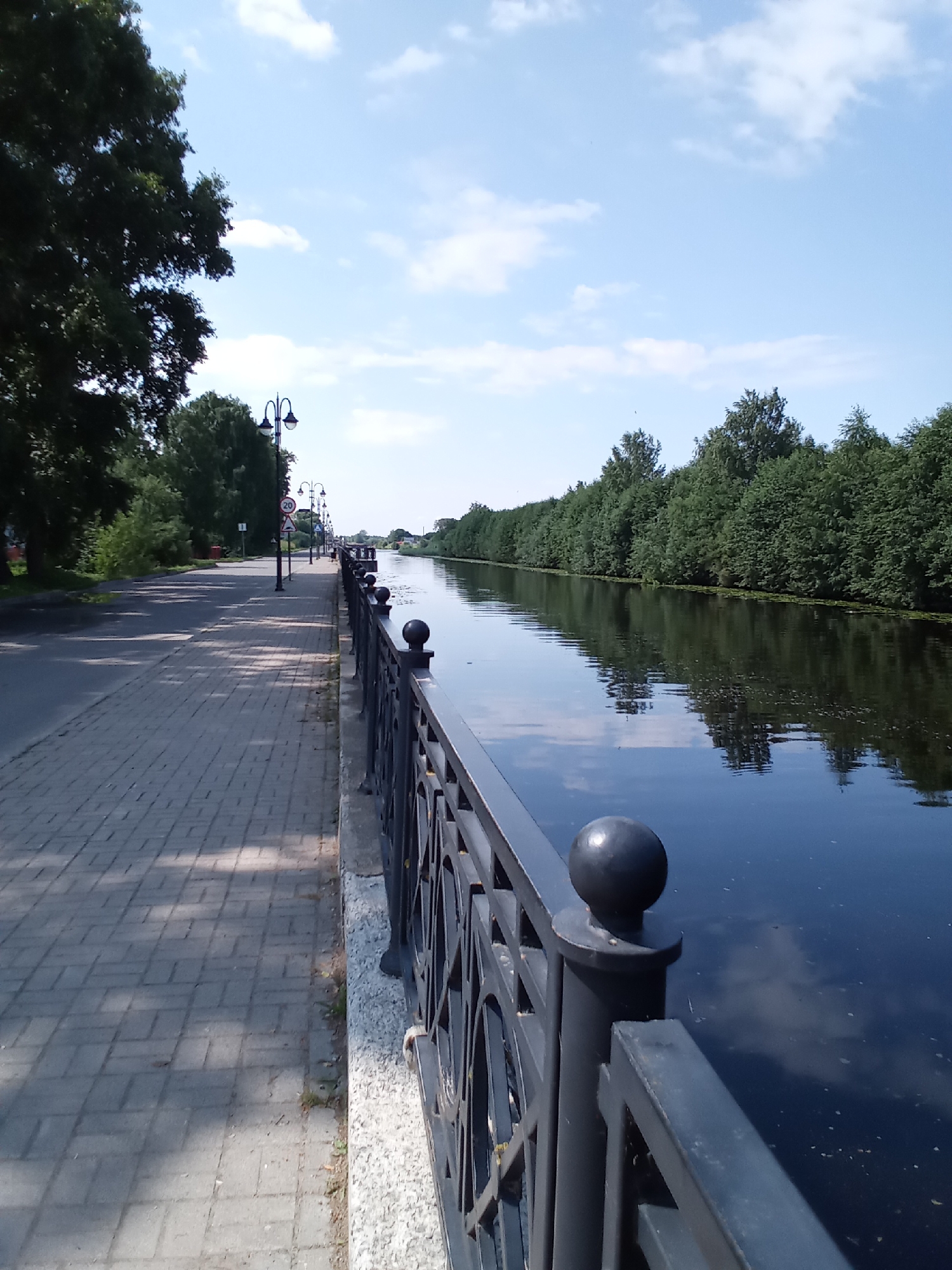 The image depicts a scenic view of a wide canal in Belozersk, Russia, on a clear day. The perspective is from a paved walkway lined with a decorative black metal railing. The walkway is constructed of light-colored brick pavers, casting distinct shadows from the trees that line the left side of the path. These trees are lush and green, with a large tree dominating the foreground on the left.
Beyond the railing, the canal's dark, reflective water stretches into the distance, mirroring the bright blue sky and scattered white clouds. Along the opposite bank of the canal, a dense, continuous line of green trees forms a verdant border. The canal is calm, with only minor ripples on the surface.
Several lampposts with a traditional design are positioned along the walkway, some with accompanying traffic signs. One sign indicates a speed limit of 20. In the far distance, hints of buildings or structures are visible, suggesting a developed area beyond the tree line. The overall atmosphere is tranquil and picturesque, characteristic of a park or riverside promenade. No people are visible in the scene.
