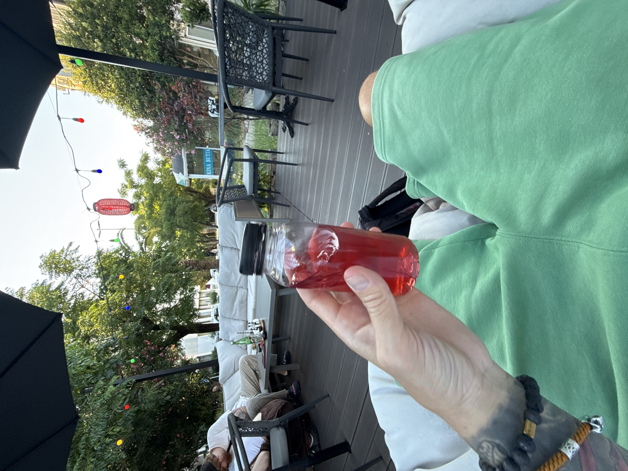 In Bar, Montenegro, an outdoor seating area features a person holding a clear plastic bottle containing a red beverage. The person is wearing green shorts and has tattoos on their arm. In the background, a group of people are seated on a white couch with a table in front of them. The area is decorated with string lights and a red lantern. The scene appears to be a cafe or restaurant patio.