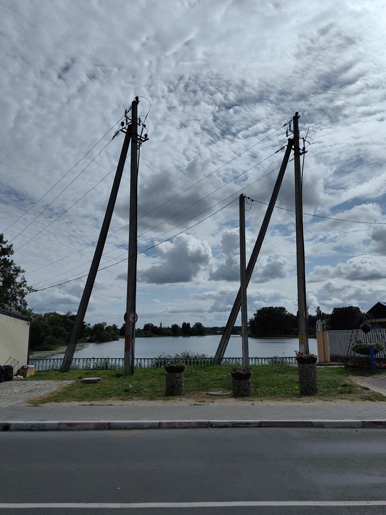 The image depicts a wide shot of a street in Pastavy, Belarus, with a body of water and trees visible in the background. The foreground features an asphalt road with a paved sidewalk. Two large, angled power poles dominate the center of the frame, supporting electrical wires that stretch across the scene. In front of the power poles, a strip of grass separates the sidewalk from a low green fence bordering the water. Two stone planters with small plants are situated on the grass. Further in the background, the water's edge is lined with trees. The sky is overcast with large, white clouds. A light-colored building with dark refuse bins is visible on the far left, and on the right, a white fence with some decorative elements and hanging baskets of flowers can be seen, adjacent to a structure. The scene appears to be a public area with infrastructure and natural elements.