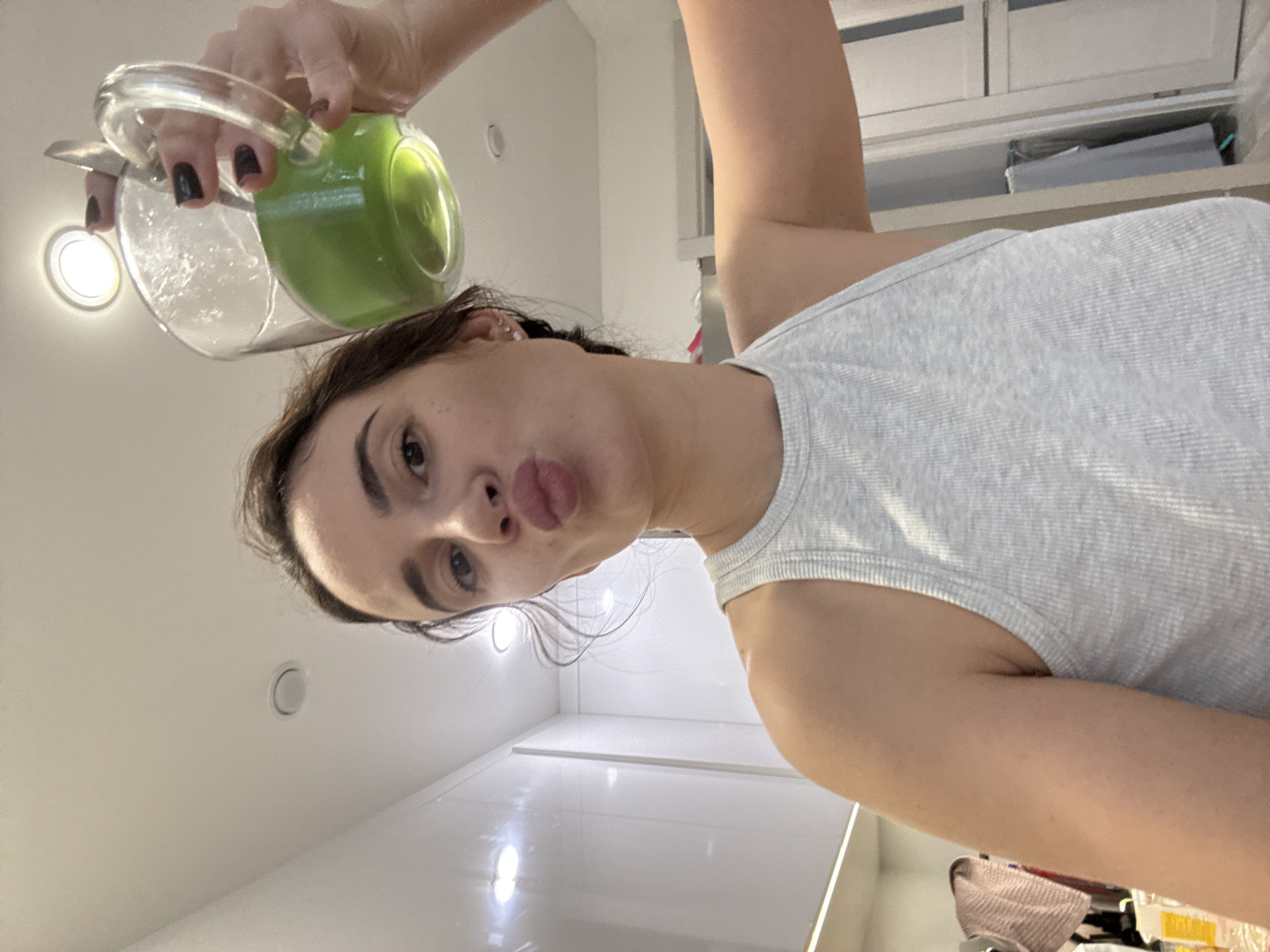 A woman in a gray tank top is seen from a low angle, holding a glass pitcher containing a green liquid over her head. The background shows a white ceiling with recessed lighting and white cabinets. The location is not identifiable as Chaikovsky, Russia from the given image.