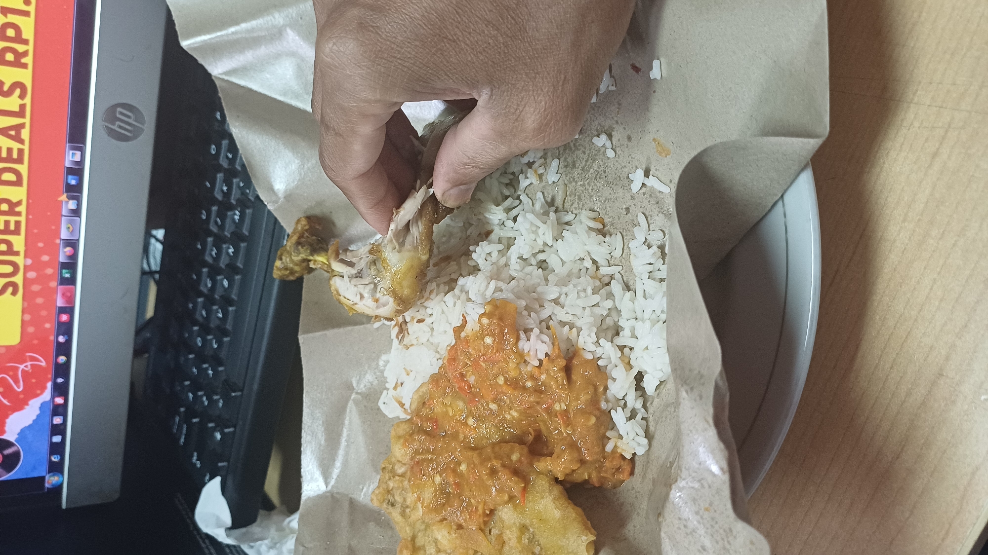 The image shows a hand holding a piece of fried chicken over a plate of white rice and what appears to be fried tempeh with chili sauce. The food is presented on a brown paper wrapper, which is resting on a white plate. In the background, a computer monitor displays a promotional image with the text "SUPER DEALS RP1." A keyboard is visible beneath the monitor. The setting appears to be a desk, likely in an office or home environment, in South Jakarta, Indonesia. The lighting is artificial.
