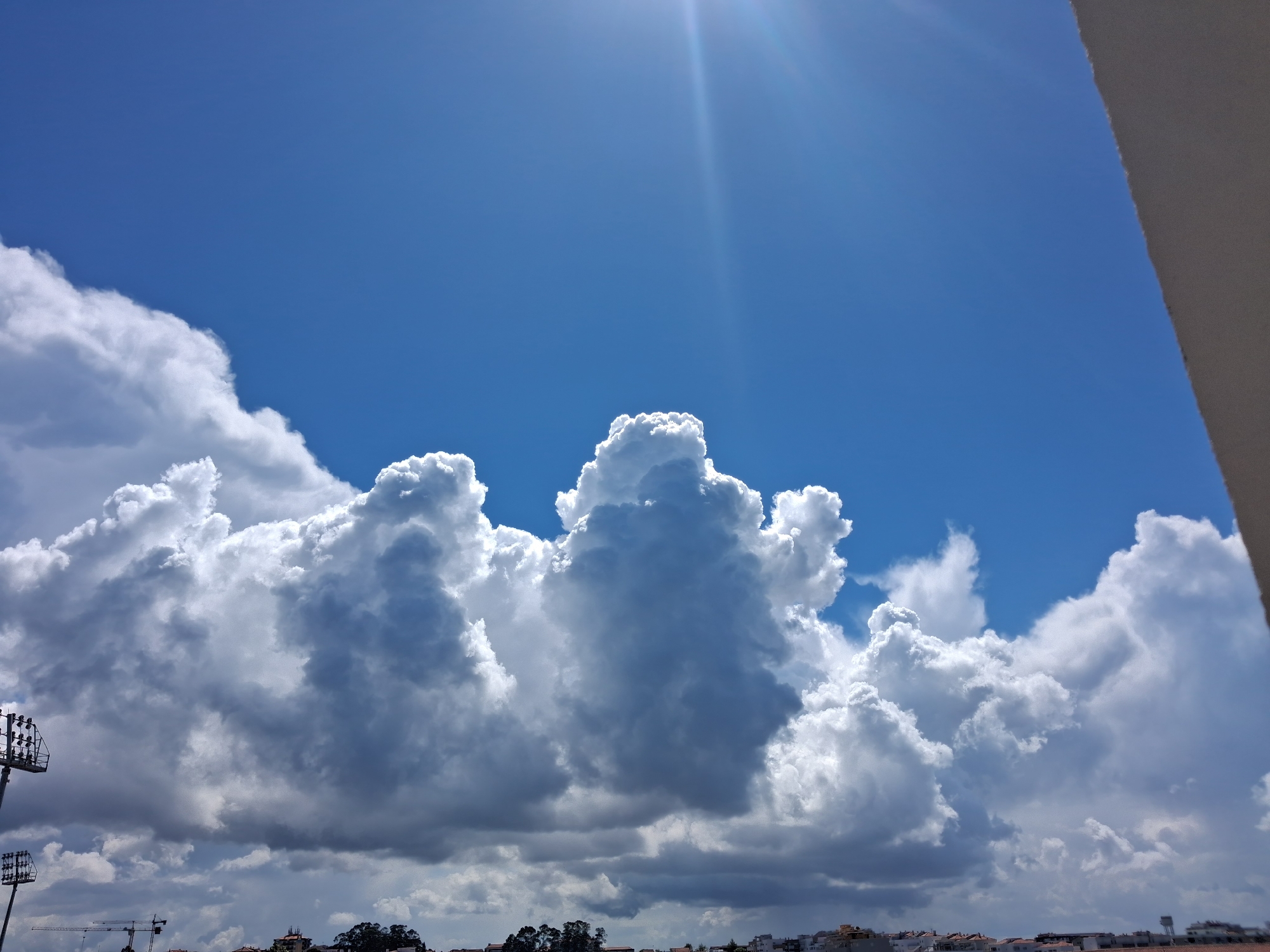 The image shows a daytime view of the sky above Figueira da Foz, Portugal. Predominantly blue, the sky is filled with large, cumulus clouds of varying shades of white and gray. The sun is visible as a bright glare in the upper portion of the image. In the lower portion, indistinct buildings and trees are present on the horizon line. A lighting support tower is to the left. A portion of a white or light-colored building is visible in the upper right corner. There are also construction cranes.