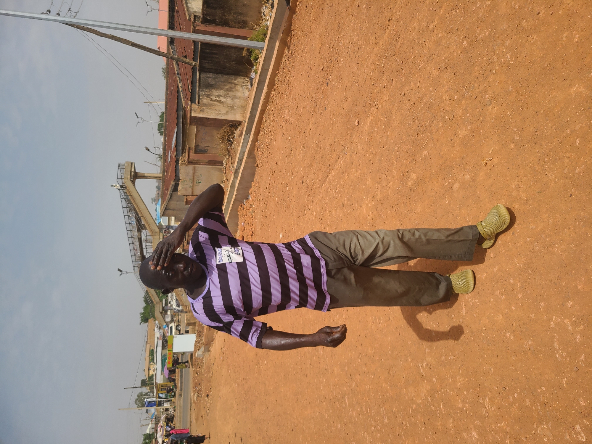An adult male stands on a dusty, reddish-brown ground surface, likely soil or unpaved road, located on the outskirts of a city identified as Jos, Nigeria. The man is wearing a purple and black striped shirt, khaki pants, and textured greenish-yellow shoes. He has his right hand on his forehead as if shading his eyes or in distress. In the background, there is a partially obscured building with a corrugated roof, various urban structures, and a glimpse of distant buildings along a horizon. The sky appears overcast.