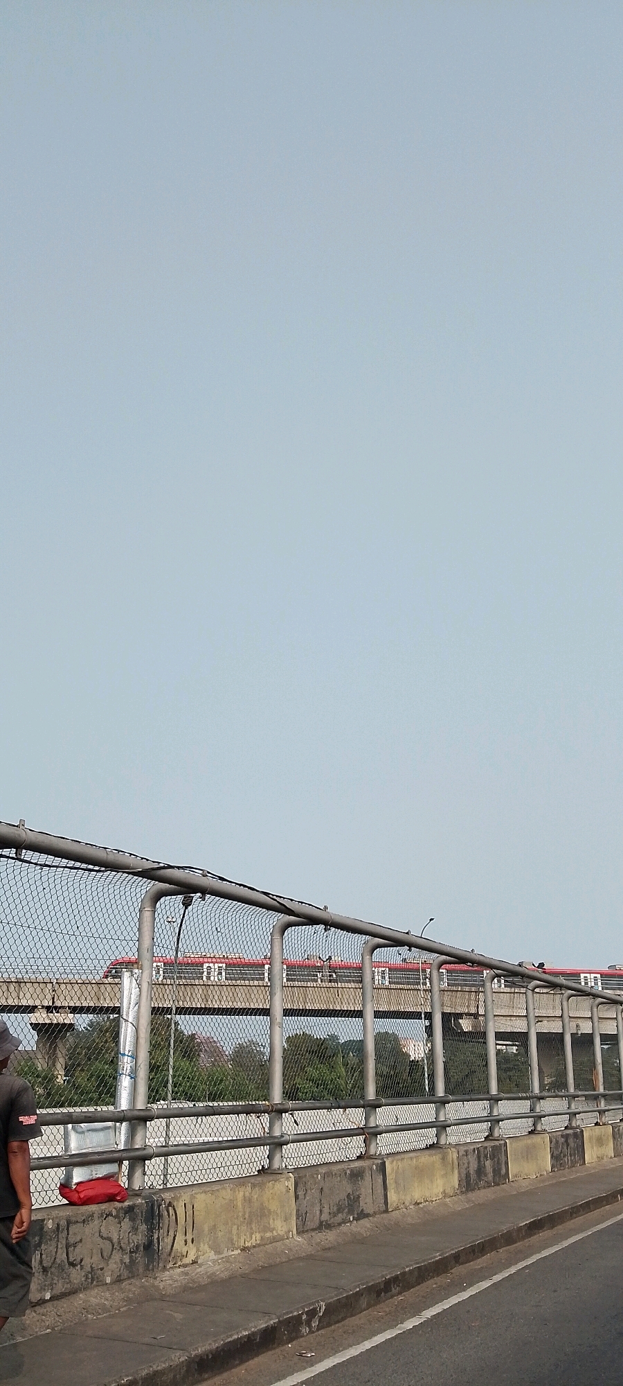 This image depicts a street-level view of an elevated train track in East Jakarta, Indonesia, on what appears to be a clear day with a pale blue sky. In the foreground, a chain-link fence with metal supports runs horizontally across the frame, partially obscuring the view of the train and its surroundings. A concrete barrier, showing signs of graffiti and weathering, runs along the bottom of the fence. To the left of the frame, the lower half of a person wearing a dark shirt and shorts is visible, walking away from the camera. On top of the concrete barrier, a red bag and a stack of white rectangular objects are placed.
The elevated train track is a prominent feature in the mid-ground, supported by concrete pillars. A red and white train with visible windows is traveling along the track. Beyond the train, a line of green trees and foliage suggests an urban landscape. The road surface is visible in the lower right portion of the image, marked with a white line. The scene conveys a typical urban environment with infrastructure for public transportation.