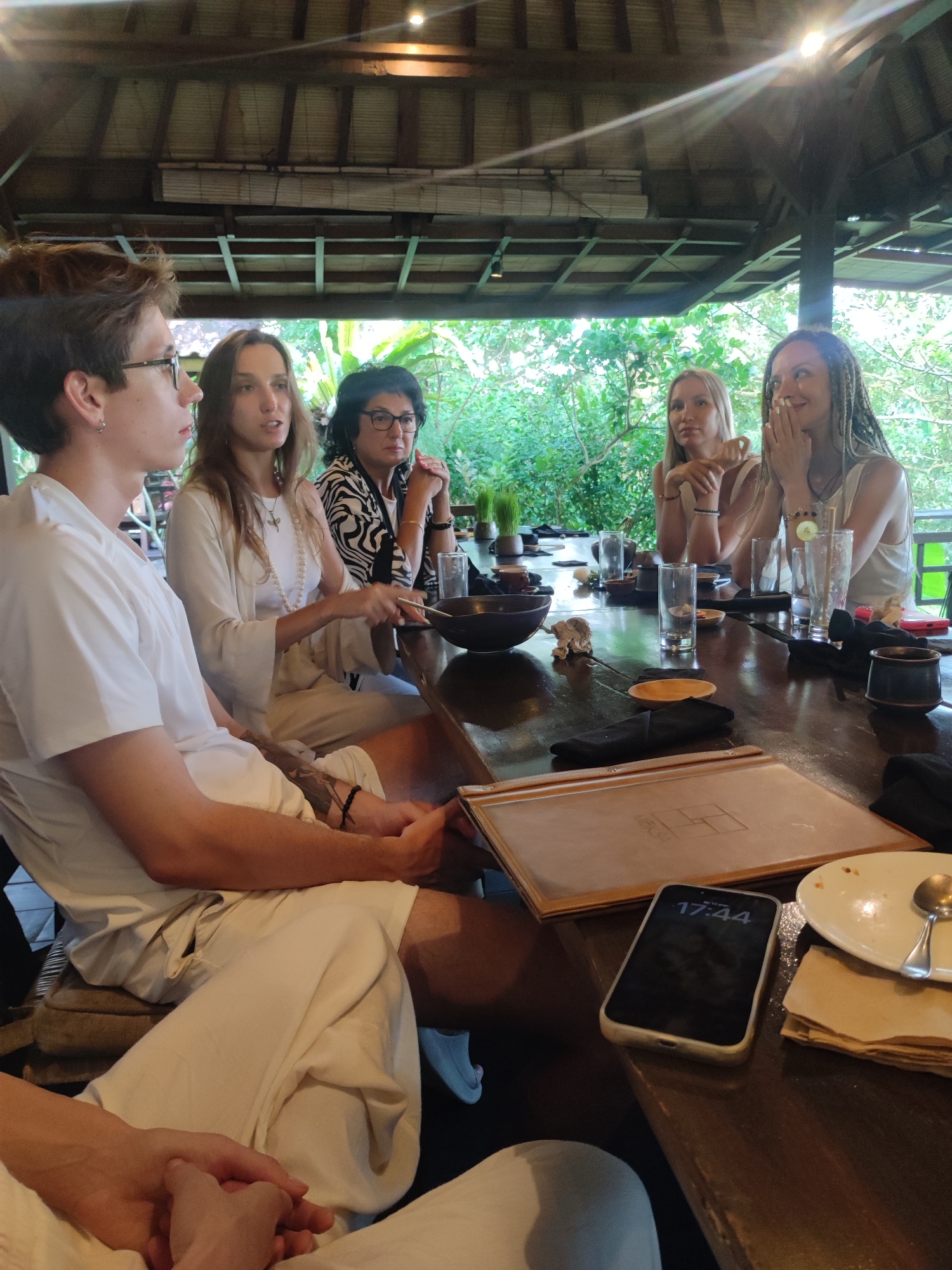 A photograph depicts a group of five people seated around a dark wooden table within an open-air structure. The location is Sayan, Indonesia.
The group consists of three women and two men. The individual nearest to the camera is a young man with brown hair, wearing a white shirt and trousers. He has glasses and an earring. Beside him sits a woman with long brown hair and a white outfit. The third individual is a woman wearing a black and white patterned top and glasses. On the opposite side of the table sit two other women, both with blond hair, wearing white tops. One has dreadlocks.
The table is covered with glasses, bowls, napkins, and a menu. A smartphone with a screen displaying "17:44" lies near the edge of the table. The setting is outdoors, with green vegetation visible in the background. The structure has a roof made of dark wood.