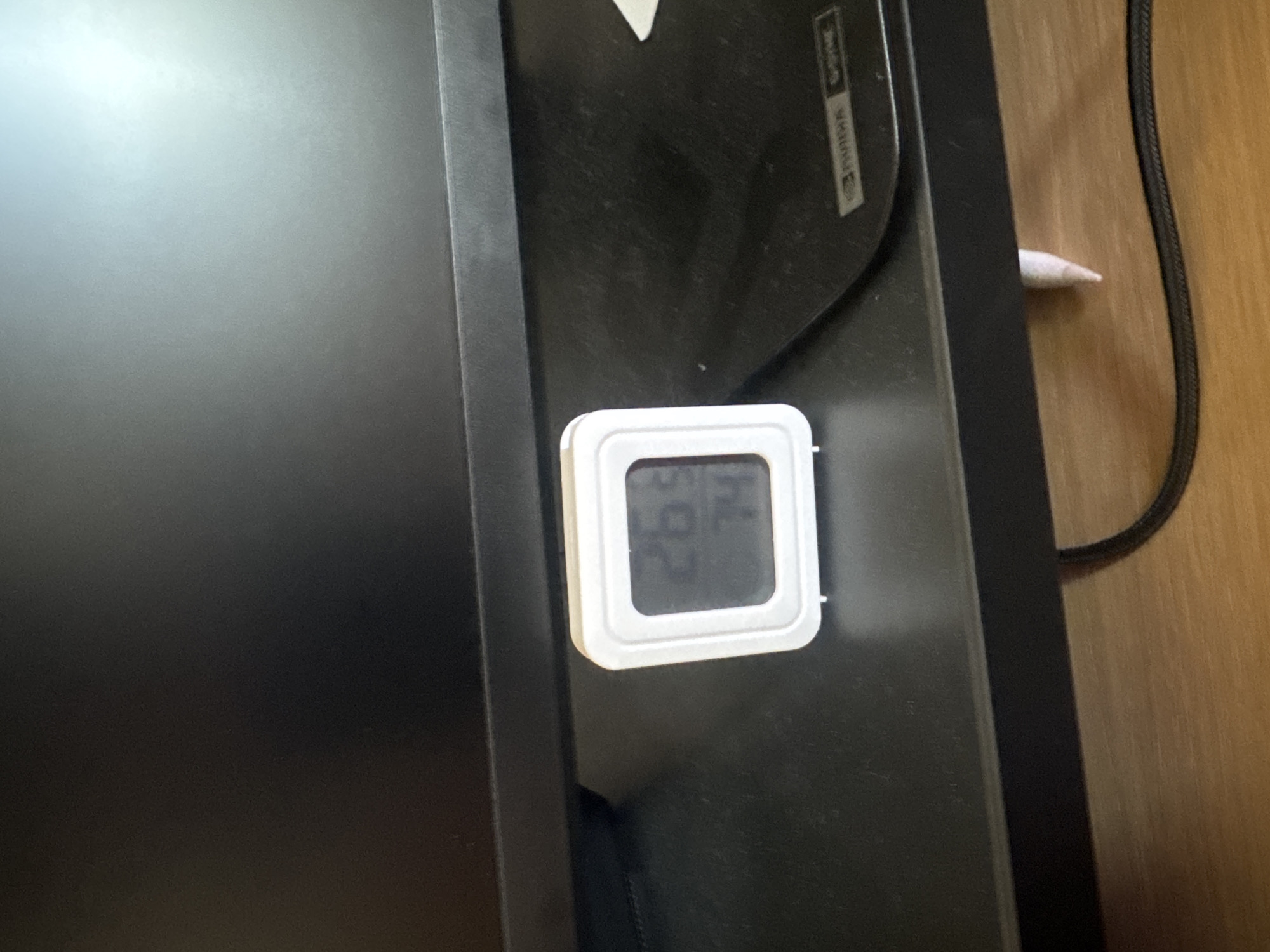 A small, white digital thermometer is attached to the black frame of a monitor. The thermometer displays "26.9" on the left side of its screen, and "74" on the right, likely representing temperature and humidity respectively. The monitor itself is dark and appears to be a computer monitor, positioned on a wooden surface. A black power cord snakes along the right edge of the frame, and a cotton swab with a pink tip is visible protruding from a gap in the monitor's housing. The background suggests an indoor setting with a wooden desk or table. The lighting is somewhat dim, with a bright glare on the left side of the monitor screen. No people are visible, and the scene is static, showing a close-up of the thermometer and part of the monitor. Text on the monitor's bezel is partially visible and appears to be a brand logo or product information, but it is too blurry to read definitively. The overall impression is a functional setup, possibly in a home or office environment in Livadia Larnakas, Cyprus.