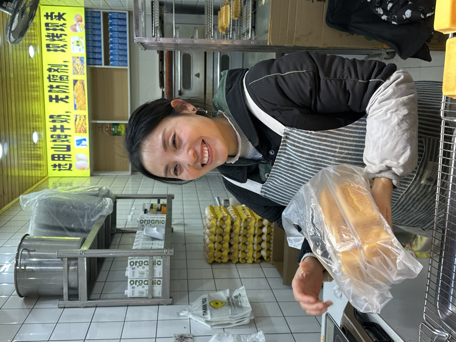 An indoor commercial bakery or food stall in Jiangjin, China, is depicted. A female, identified as an employee, is positioned centrally, smiling broadly with her head turned slightly towards the right. She has dark hair and is wearing a black padded jacket, a striped apron (white and gray), and light gray arm sleeves. In her hands, she holds a clear plastic bag containing four golden-brown rectangular loaves of baked goods, likely bread or cake.
The background features white tiled walls with gray grout. To the left, a two-tiered stainless steel shelf holds a large lidded pot and several plastic-wrapped items on the upper shelf, and multiple "organic" branded cartons on the lower shelf. Below these shelves, a tall stack of yellow egg trays containing white eggs is visible. A white plastic "THANK YOU" bag hangs on the wall.
To the right of the woman, a commercial oven or display unit with multiple racks contains additional golden-brown rectangular loaves. Above this, a yellow sign with red and black Chinese characters translates to "Selected Sam's Club Milk, no preservatives, freshly baked and sold," and includes illustrations of eggs and baked goods. Below the sign, shelves hold blue-labeled cartons and a bottle of cooking oil. A ceiling fan is partially visible in the upper right. The foreground includes a stainless steel counter with a metal grate, and more unwrapped baked loaves are visible on the far right. The scene is brightly lit.