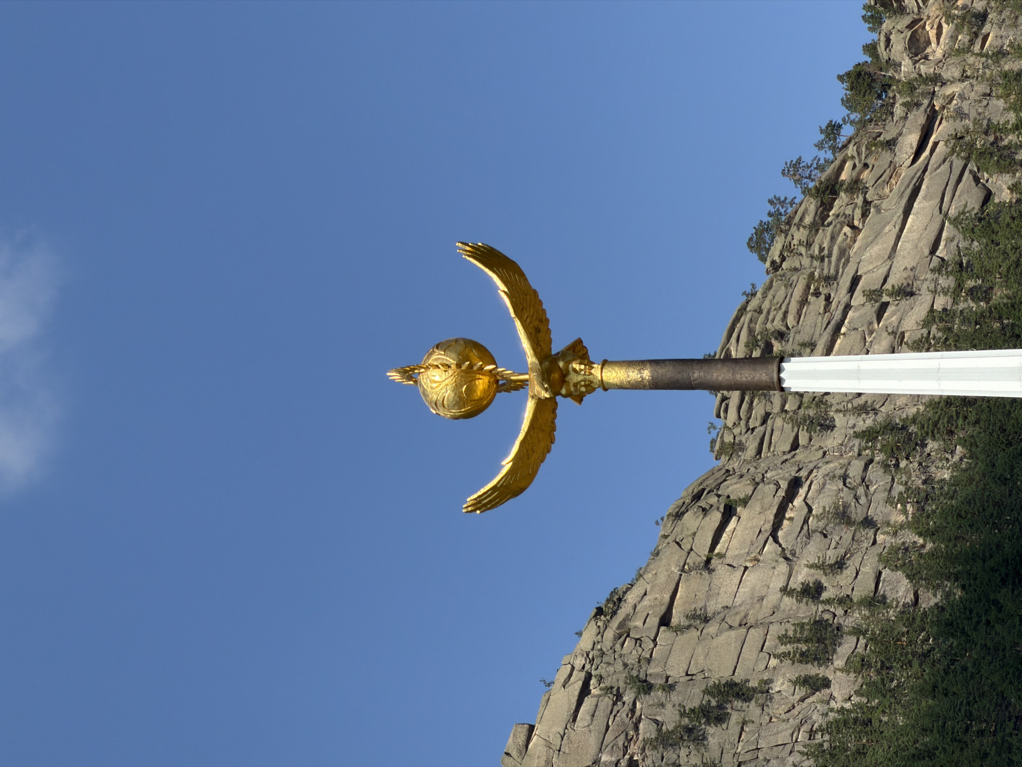 The image shows a golden sculpture of a bird with outstretched wings holding a globe on top of a pole. The sculpture is positioned in front of a rocky mountain range with sparse trees growing on it. The sky is clear and blue with a few white clouds. The sculpture is made of a shiny, metallic material and appears to be a significant landmark. The rocky formations are layered and weathered, suggesting an arid or mountainous environment. The trees are coniferous and appear to be growing in crevices and on the slopes of the rocks. The overall scene conveys a sense of grandeur and natural beauty. This landmark is located in the village of Zelenyy Bor, in the Burabay district of Akmola region, Kazakhstan, a location known for its stunning natural landscapes and unique rock formations.