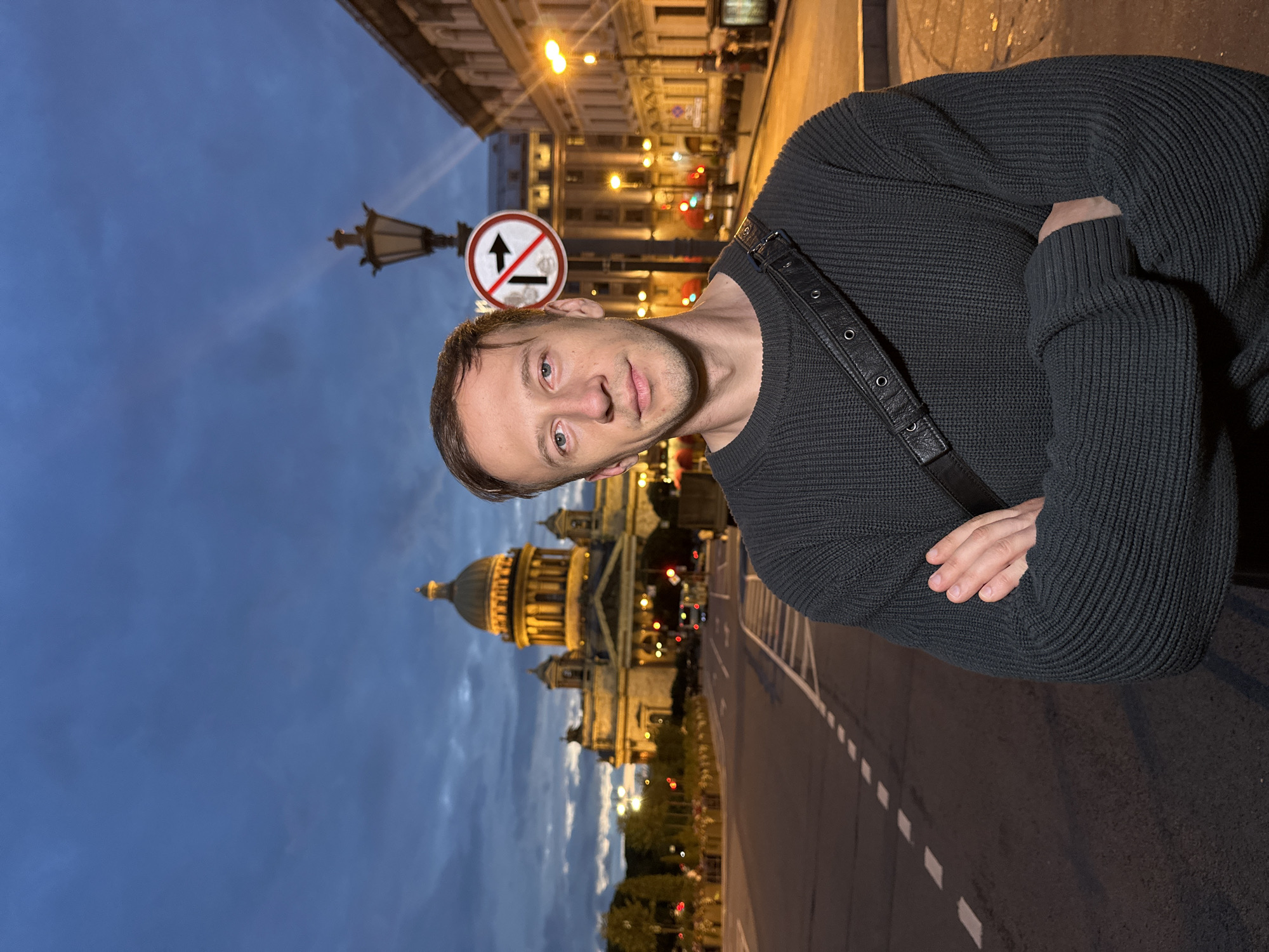 A man stands with his arms crossed in front of Isaac's Cathedral in Saint Petersburg, Russia. The sky is a deep blue, indicating dusk or early night. The cathedral is illuminated by warm yellow lights, highlighting its dome and architectural details. Streetlights and building lights line the wide road in the foreground, casting a glow on the asphalt. A traffic sign prohibiting a right turn is visible on a lamppost to the right of the man. The man is wearing a dark, ribbed sweater and a black strap across his chest. The scene is set on a paved street with white dashed lines marking lanes.