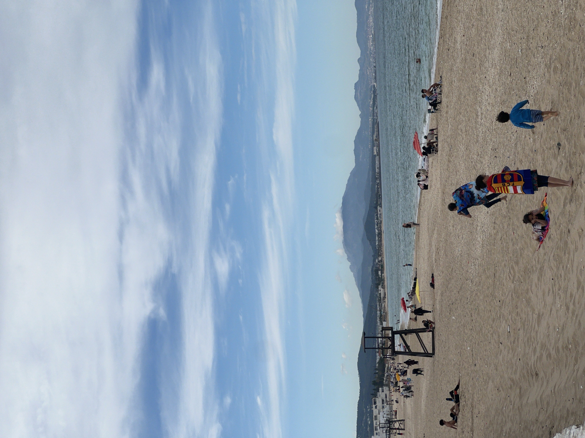 The image captures a coastal scene at M'Diq, Morocco, showcasing a beach populated with people. On the sandy beach, several individuals are visible, including some lying down, others standing, and one running. Adjacent to the beach is the Mediterranean Sea, with a coastline stretching into the distance. In the background, mountainous terrain adds depth to the scene. The sky above is partially covered with clouds. Beach umbrellas and what appear to be lifeguard structures are also visible along the shoreline.