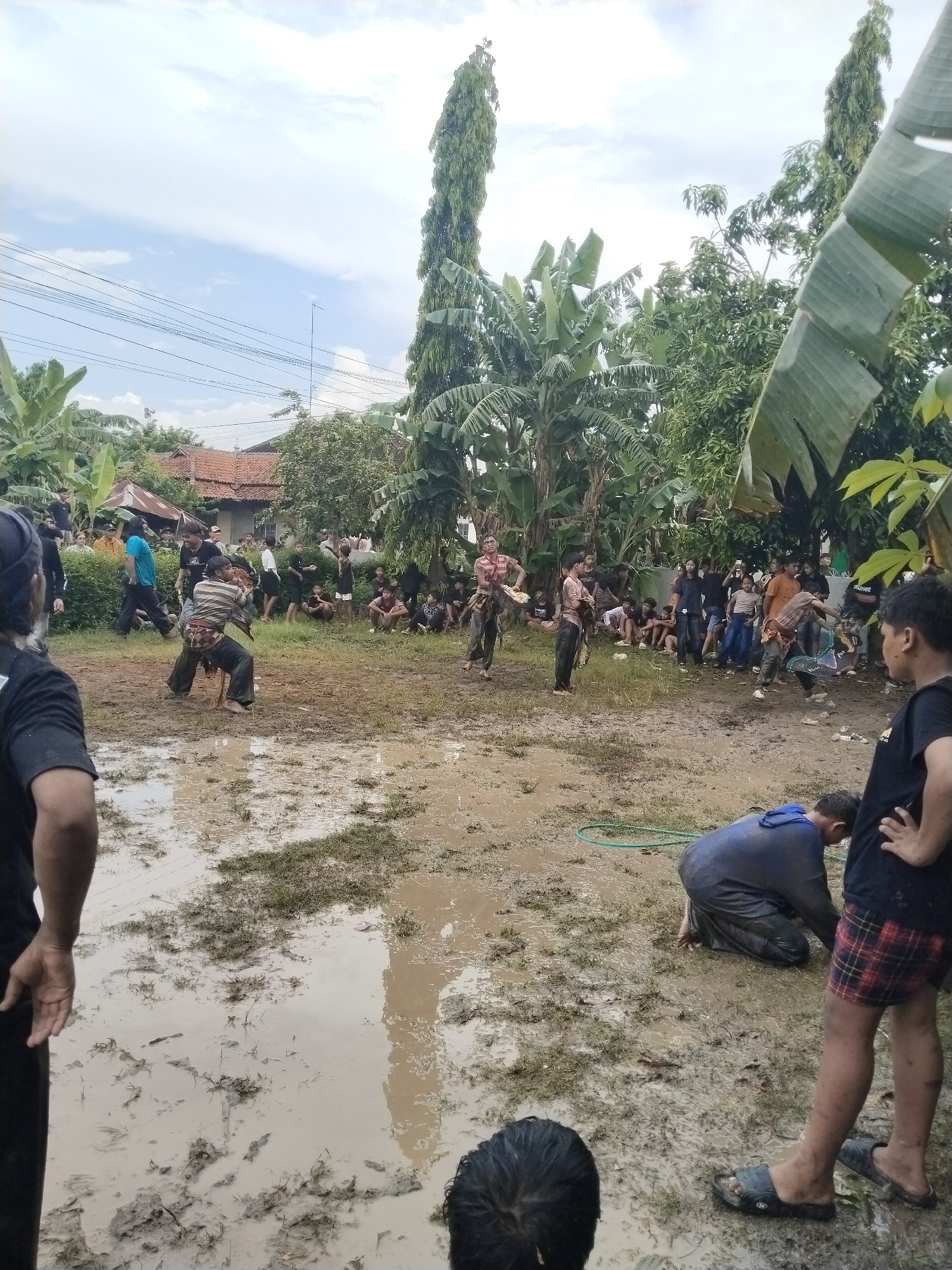 The image depicts a muddy area in Karang Tengah, Indonesia, where a crowd has gathered. In the center, two individuals are engaged in a physical activity, possibly a traditional fight or game. The muddy ground suggests recent rain. Several people are observing the event, while others are partially submerged in the mud. The surrounding area includes houses and dense tropical foliage, with banana trees prominently visible. Overhead power lines are strung between poles. The overall atmosphere indicates a local gathering or event.