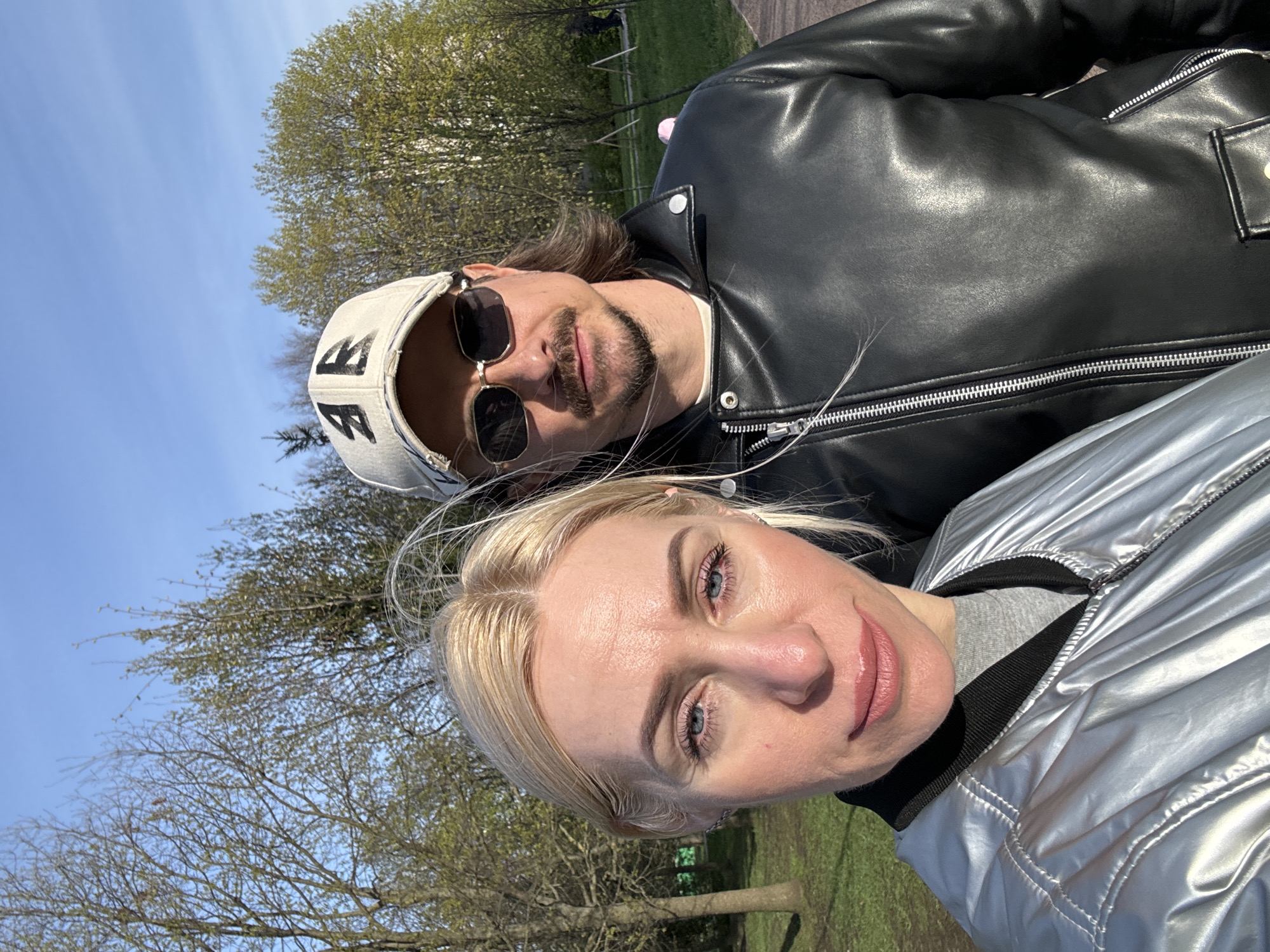 A woman with blonde hair wearing a silver jacket and a man with long brown hair wearing sunglasses, a baseball cap, and a black leather jacket pose for a selfie. They are outside, in what appears to be a park. Trees with new spring growth are visible in the background, along with blue sky. The location may be Saint Petersburg, Russia.
