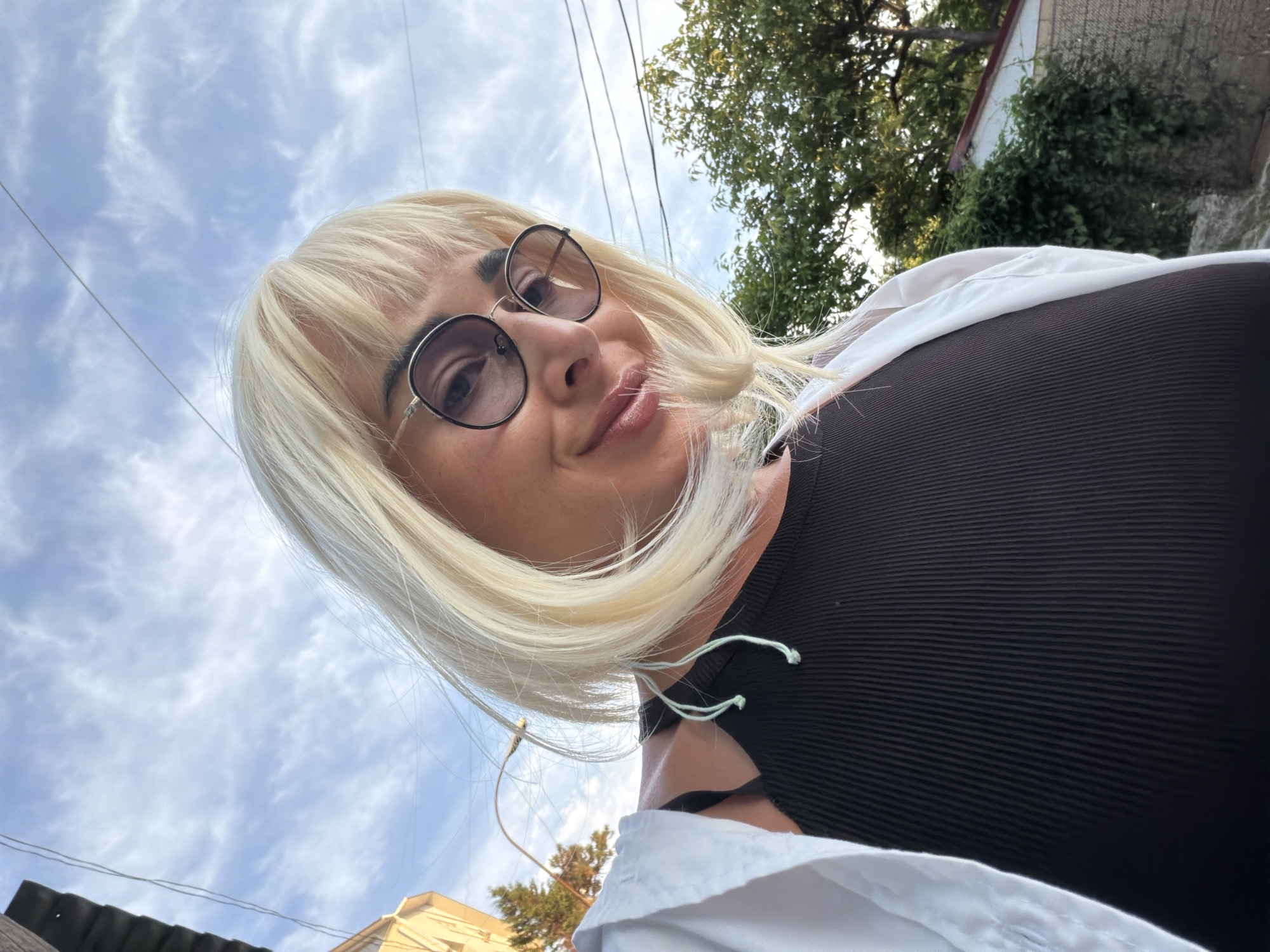 A blonde woman wearing glasses and a black ribbed top, with a white shirt draped over her shoulders, is captured in a low-angle selfie. She has full lips with a pinkish-brown lipstick and her bangs frame her face. The sky, with scattered white clouds, fills much of the background, suggesting an outdoor setting. A few power lines are visible against the sky. Trees and the upper part of a building can be seen to the right and in the distance, indicating a somewhat urban or suburban environment. The lighting suggests it is daytime, likely late afternoon given the warm tone of the light. The woman's expression appears relaxed and natural. There is no visible text in the image. The location context is Sochi, Russia.