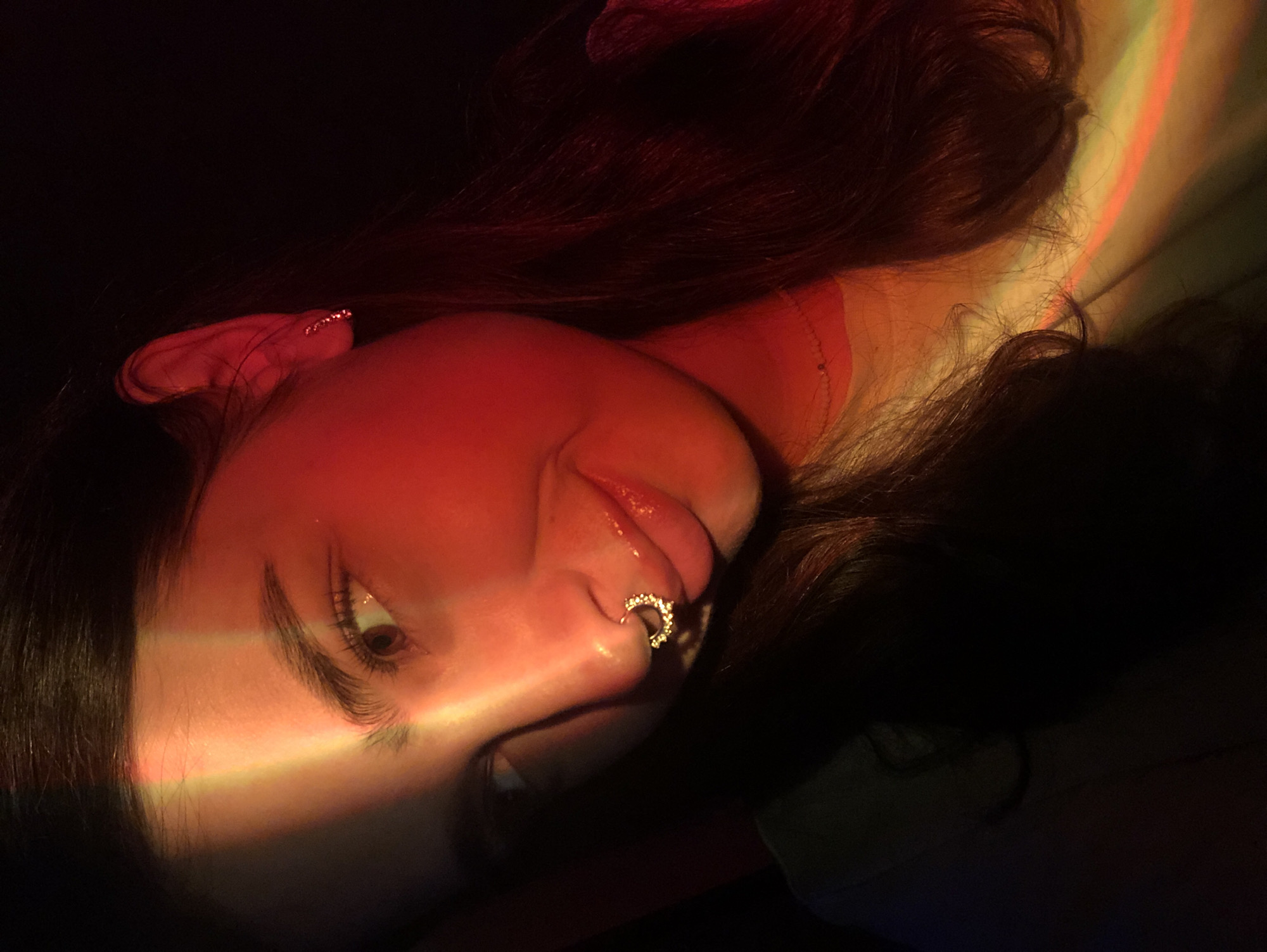 A close-up, waist-up, indoor, dimly lit selfie shows a young woman with long, dark hair, angled to her left, with her right eye and part of her forehead illuminated by a striped rainbow light effect. She has thick, dark eyebrows, a nose ring with a circular pattern of small, clear stones, and full, glossy lips with a subtle smile. Her left ear, adorned with a small, delicate earring, is visible. The lighting casts a strong red hue on the right side of her face and hair, contrasting with the shadows. The background is predominantly dark, with hints of colored light reflected on her clothing and hair. There are no discernible city features or references to Noviy, Russia.