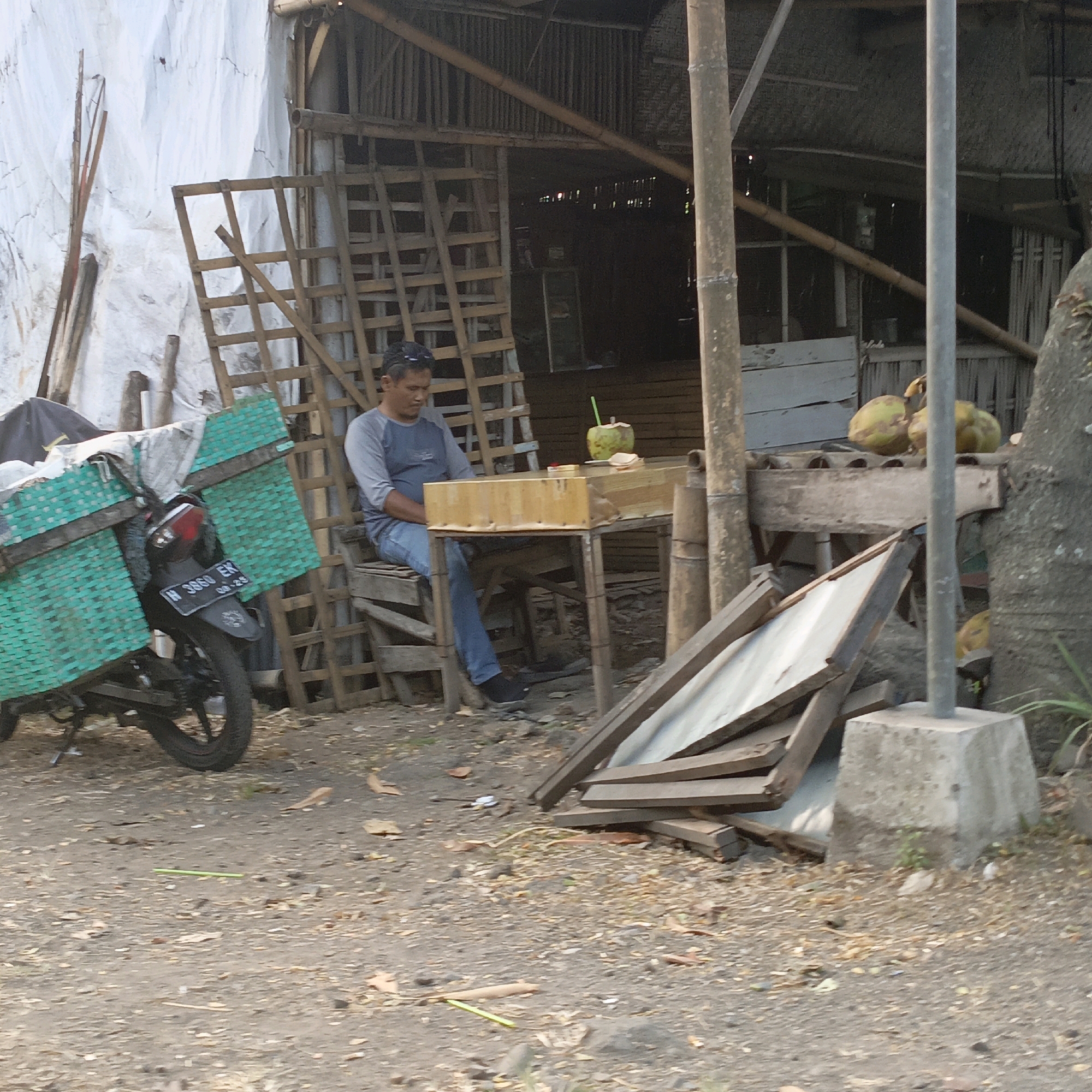 A man sits on a wooden bench at a low wooden table in front of a rustic wooden shelter. He is wearing a gray long-sleeved shirt and blue jeans. On the table are two coconuts, one with a green straw, and a pack of cigarettes. To the left of the man is a motorcycle with large green woven baskets on the back. The shelter is made of wood and bamboo, with corrugated metal roofing and a woven bamboo wall. The ground is dirt and covered with fallen leaves. In the background, there is a white tarp covering something and stacked wooden planks. This scene is consistent with a roadside vendor or rest stop in Salatiga, Indonesia.