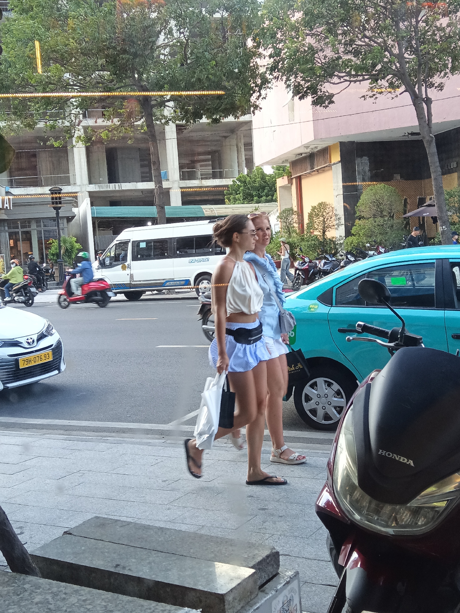 The image depicts a street scene in Nha Trang, Vietnam, focusing on a pedestrian walkway and a busy road. Two young women, dressed in light-colored summer attire, are walking side-by-side on the sidewalk, seemingly engaged in conversation. The woman on the left wears a white crop top and a light blue skirt, while the woman on the right is in a light blue dress. Both are carrying bags. They are walking towards the viewer's left, with the woman on the left in the foreground.
The street is active with traffic. A white van is visible on the road, along with several motorbikes, one of which is red and carrying a rider with a green helmet. A white car with a yellow license plate is parked on the left side of the road. A turquoise car is parked on the right side of the road, with its rear section visible. The foreground shows a motorbike, partially obscuring the view of the street and the pedestrian area. The background features buildings under construction and older structures, suggesting an urban environment undergoing development. Trees with lush green foliage line the street, providing shade. The overall lighting suggests it is daytime, possibly late afternoon, given the quality of light and the shadows.