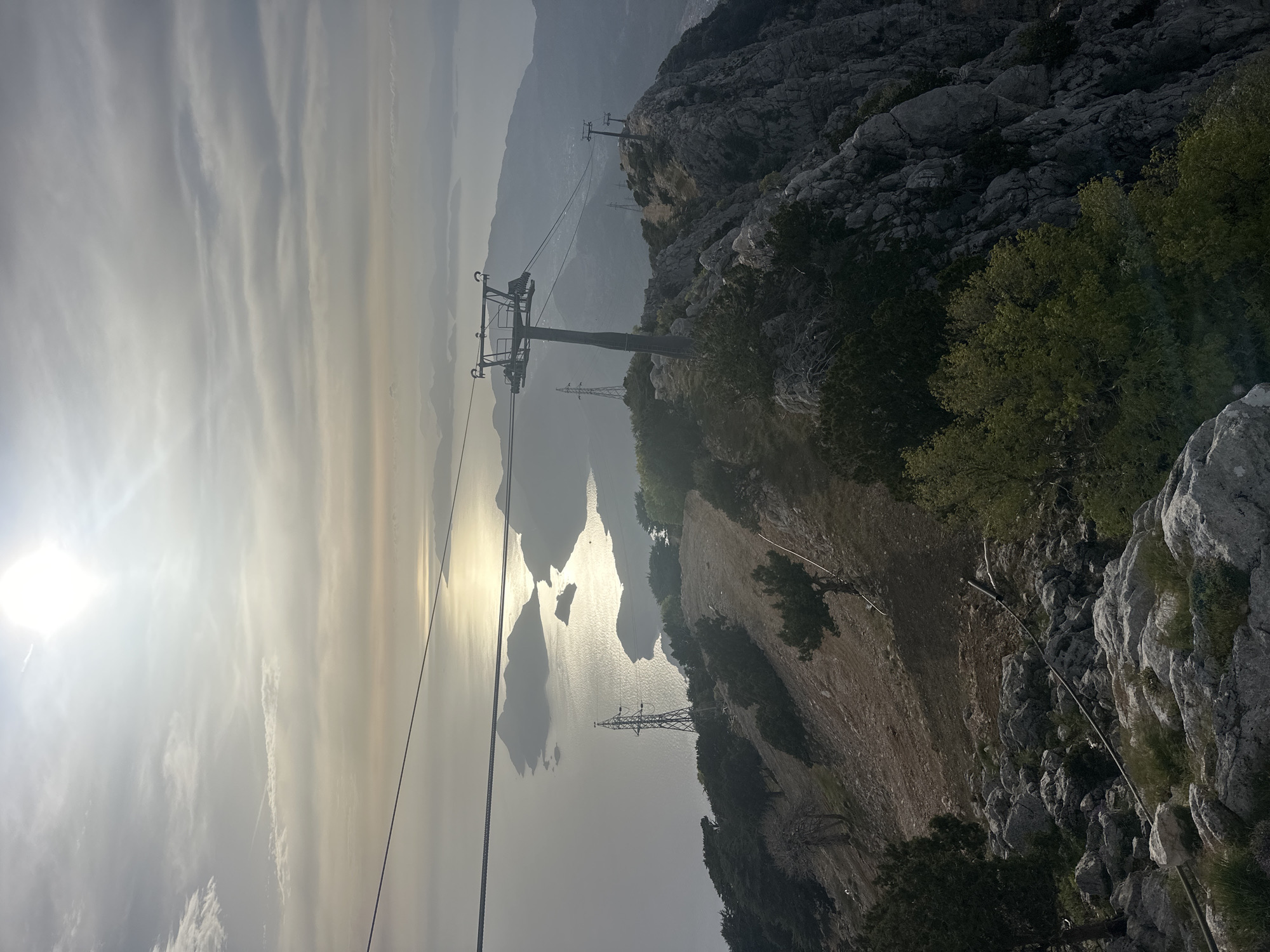 A view from a mountain overlooking the sea, possibly near Fethiye, Turkey, is captured in a vertical format. The sun glares brightly on the left, with clouds partially obscuring it. The sea reflects sunlight, and islands or landmasses appear in the distance. A cable car system's support tower and cables are visible, stretching from the foreground towards the background. An electrical power tower stands on the mountainside closer to the sea. The terrain features rocky outcrops, trees, and what appears to be a slope or cliff. A white cable is present along the ground, possibly related to utilities or infrastructure.
