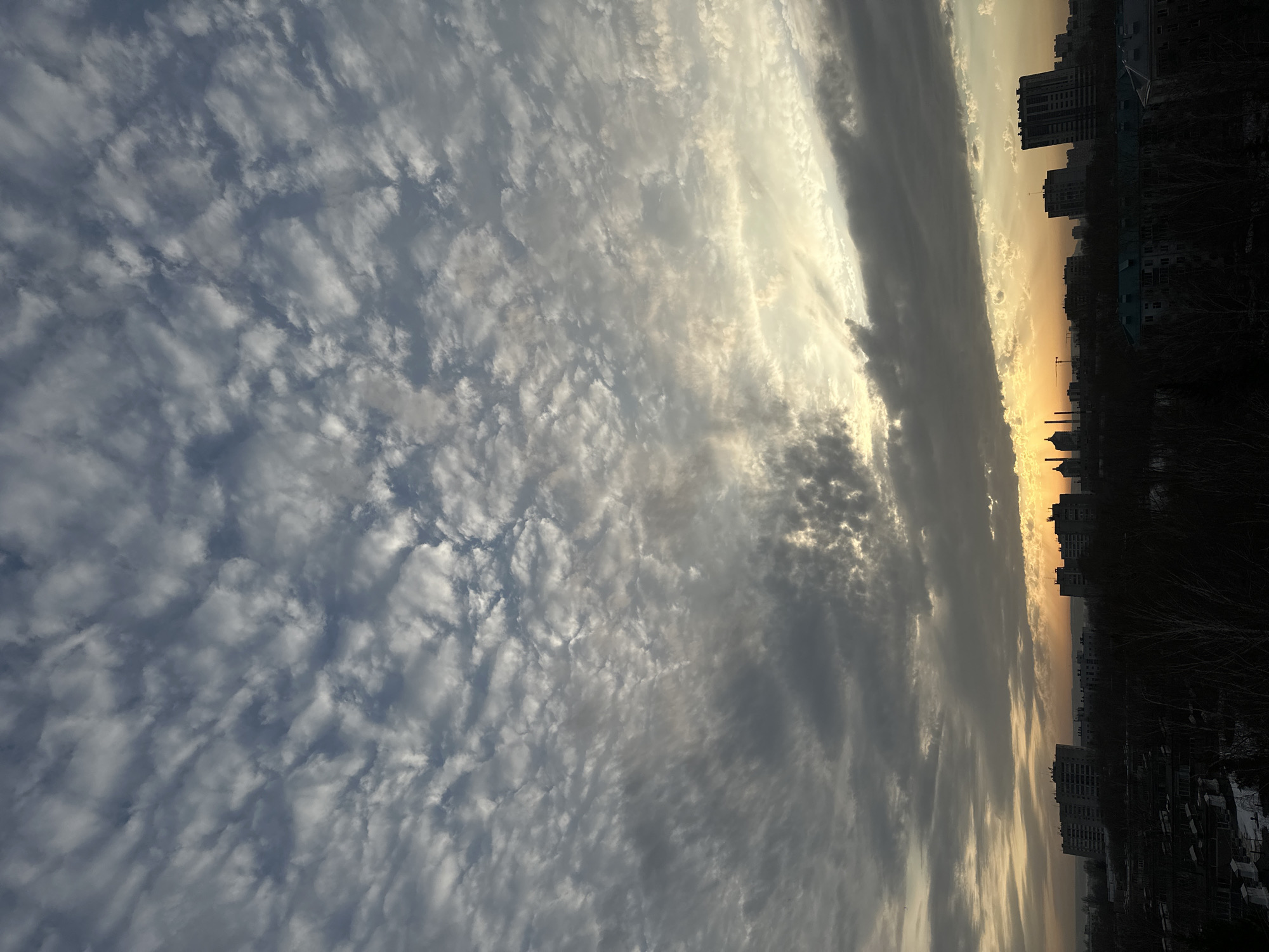 The image captures a cityscape at dusk in Kazan, Russia. The sky dominates the view, displaying a mottled pattern of clouds stretching from the lower left to the upper right, illuminated with a mix of gray and white hues. A band of darker, thicker clouds runs vertically along the right side of the frame, partially obscuring the setting sun. Below the clouds, the silhouette of Kazan's skyline emerges, punctuated by high-rise buildings and the distinctive dome of a mosque or cathedral. The lower portion of the image is predominantly dark, suggesting a ground-level view overlooking the city.