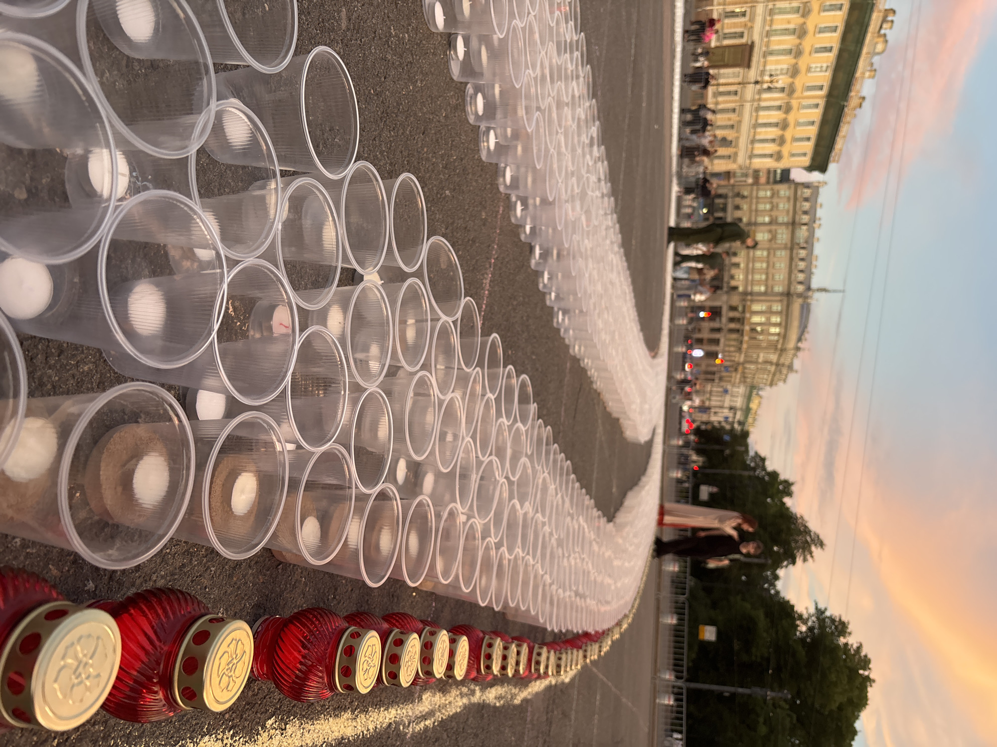 The image captures an outdoor scene at dusk in Saint Petersburg, Russia, likely on Palace Square. A vast arrangement of clear plastic cups, each containing a small white candle and sand, are laid out in a curved line across the asphalt. Adjacent to the cups, a row of red glass votive candle holders with gold lids is also displayed. In the background, several people are visible, including two individuals dressed in historical-style attire standing near the arrangement. The architecture of classical buildings, characteristic of Saint Petersburg, lines the background, under a sky painted with soft, colorful hues of sunset. The overall impression is of a solemn public installation or memorial.